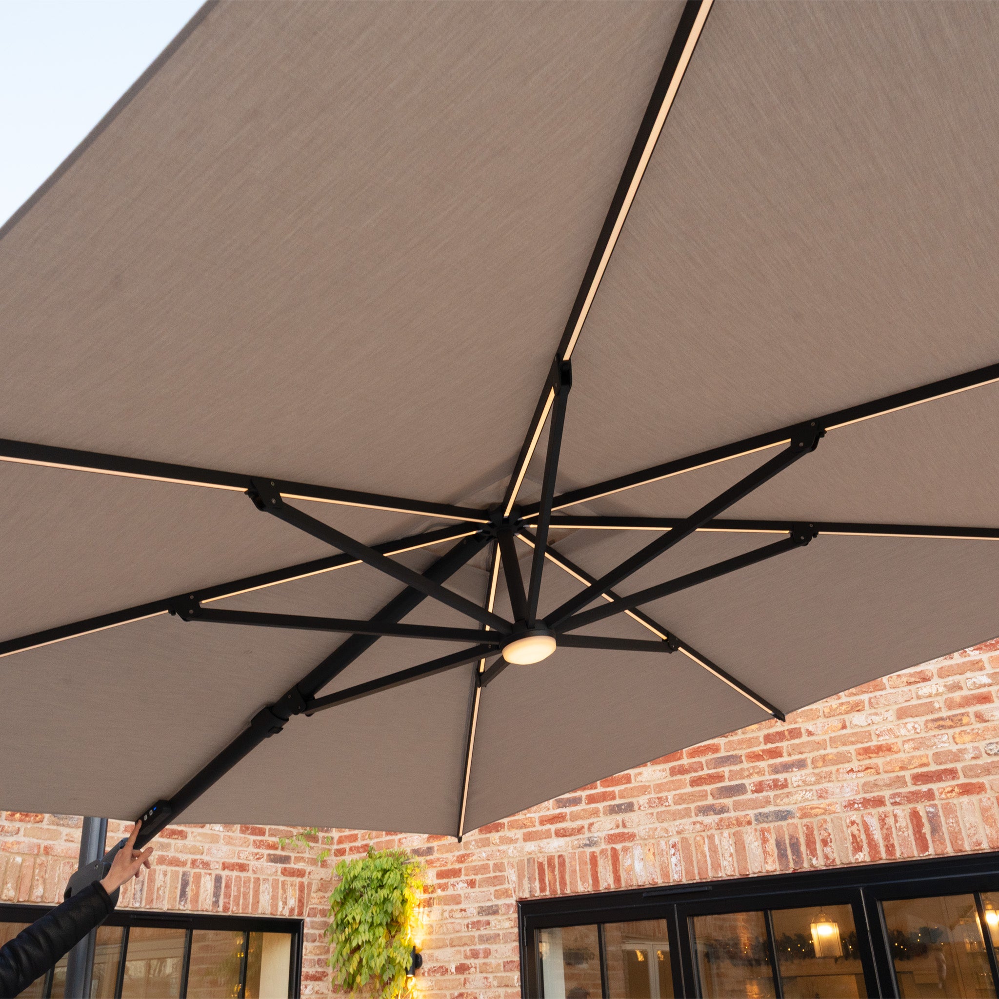 The Volos 3m x 3m Square Cantilever Parasol with LED Lighting and 120kg Granite Base in Wheat stands above a brick building, offering UV resistance and built-in LEDs with a stylish wheat canopy.