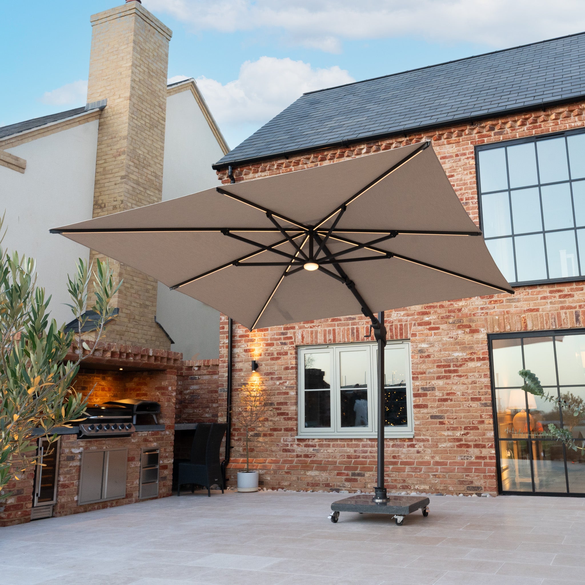 The Volos 3m x 3m Square Cantilever Parasol with LED Lighting and a 120kg granite base in Wheat provides UV protection for patios with features like brick walls, barbecue grills, and potted plants.