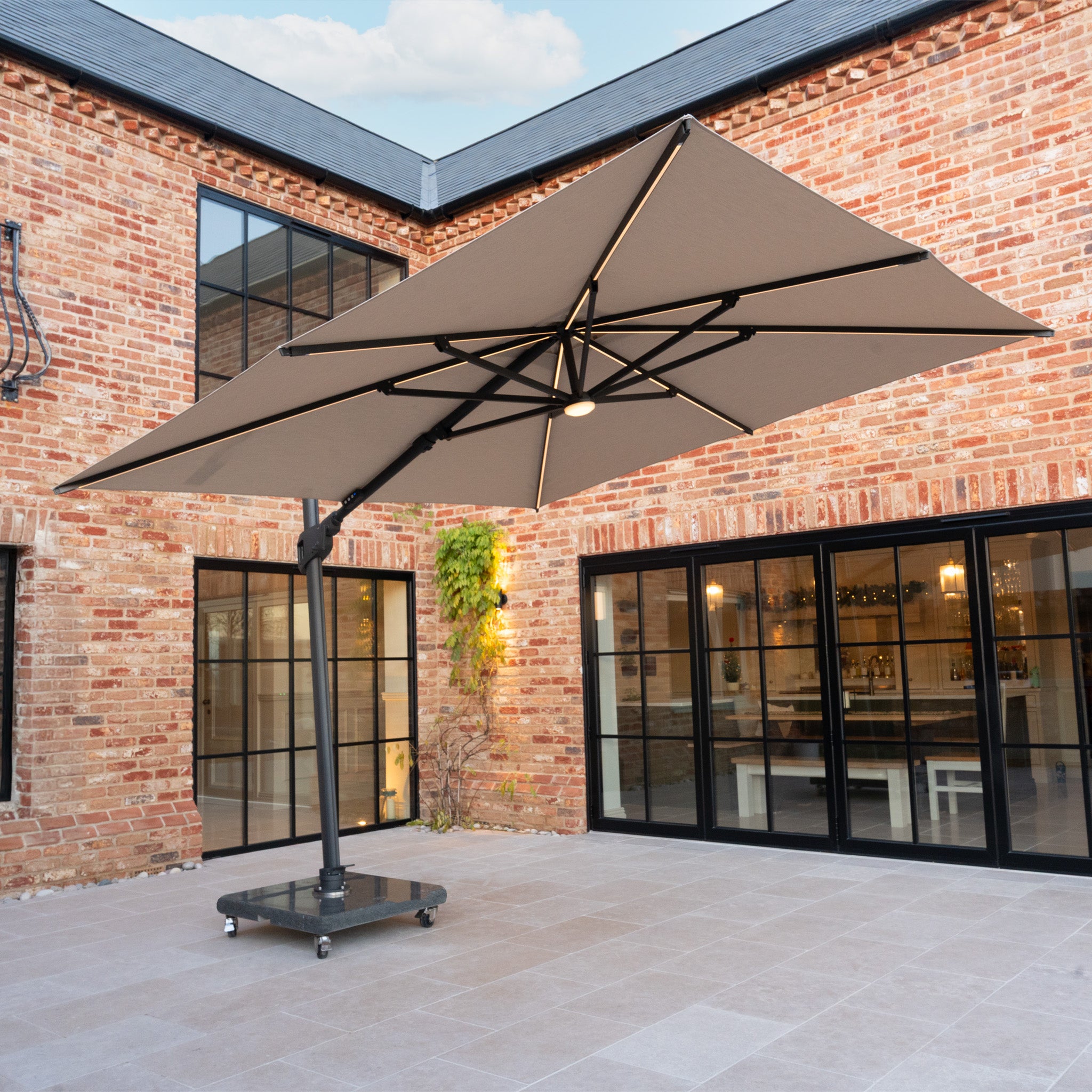 The Volos 3m x 3m Square Cantilever Parasol in Wheat with LED Lighting and a 120kg granite base stands in the courtyard, its UV-resistant canopy offering ample shade.