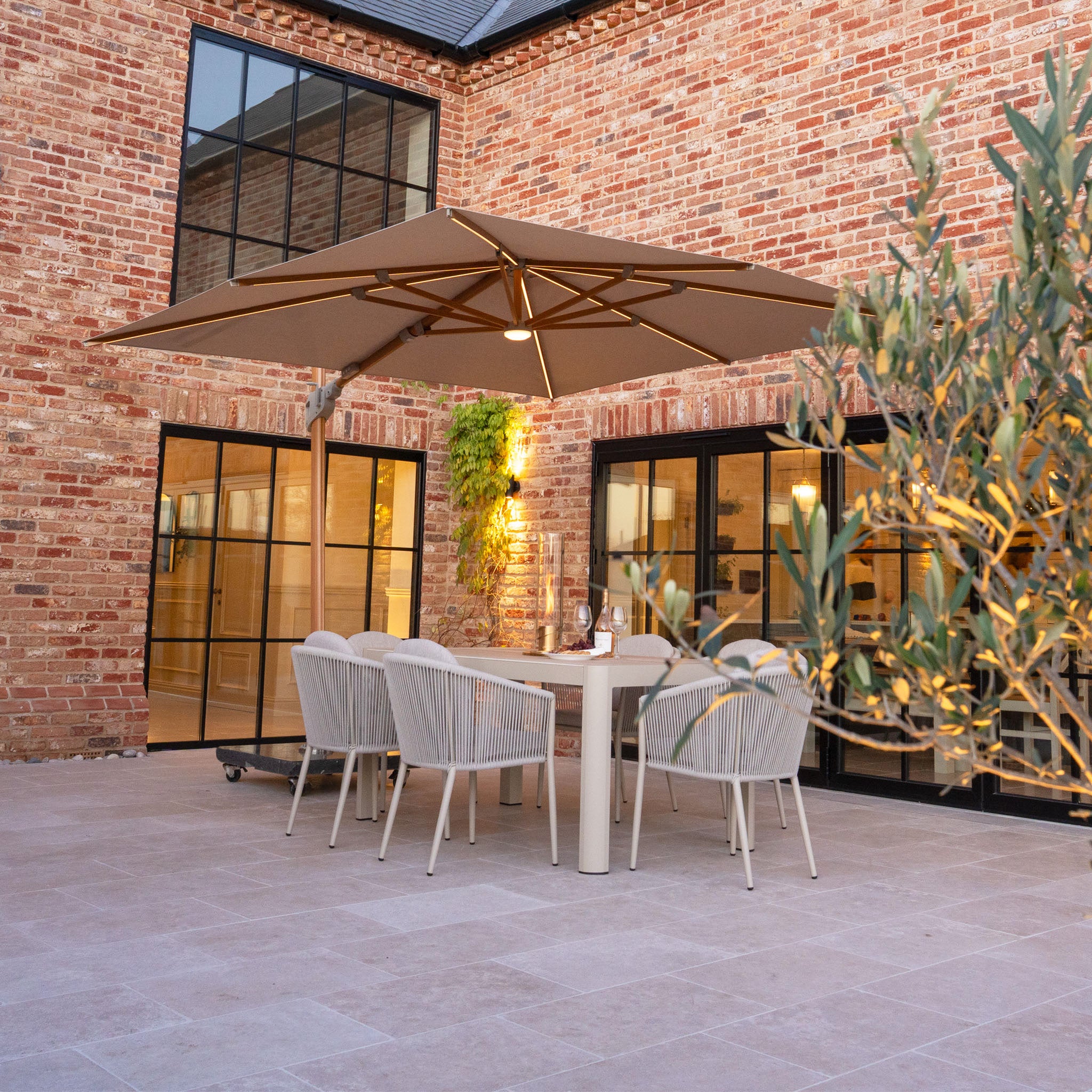 Modern patio with the Volos 3m x 3m Square Cantilever Parasol featuring LED lighting, a UV resistant wheat canopy, wood-effect finish, and a 120kg granite base, set beside a brick building with large windows.