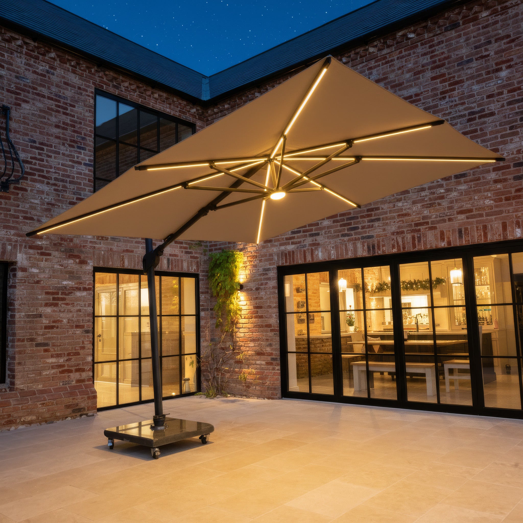 The Volos 3m x 3m Square Cantilever Parasol with LED Lighting and 120kg Granite Base in Wheat stands illuminated at night in a brick courtyard, positioned next to glass doors.
