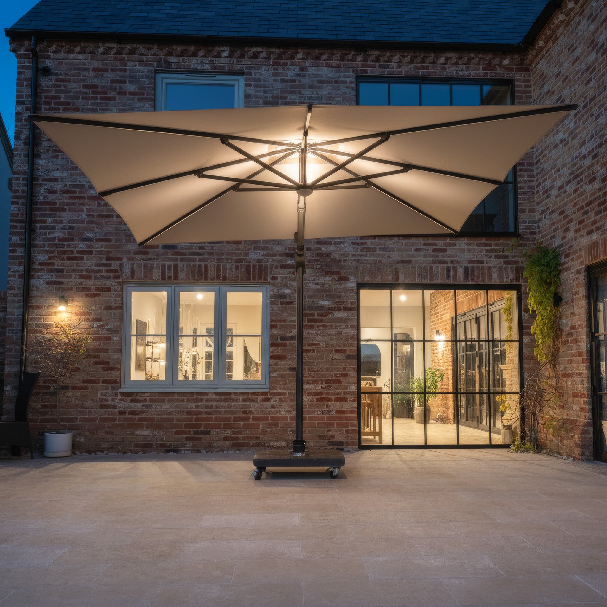 The Volos 3m x 4m Rectangular Cantilever Parasol with LED lighting and a 120kg granite base in Slate stands on the patio at dusk, illuminating the modern brick house with large windows.