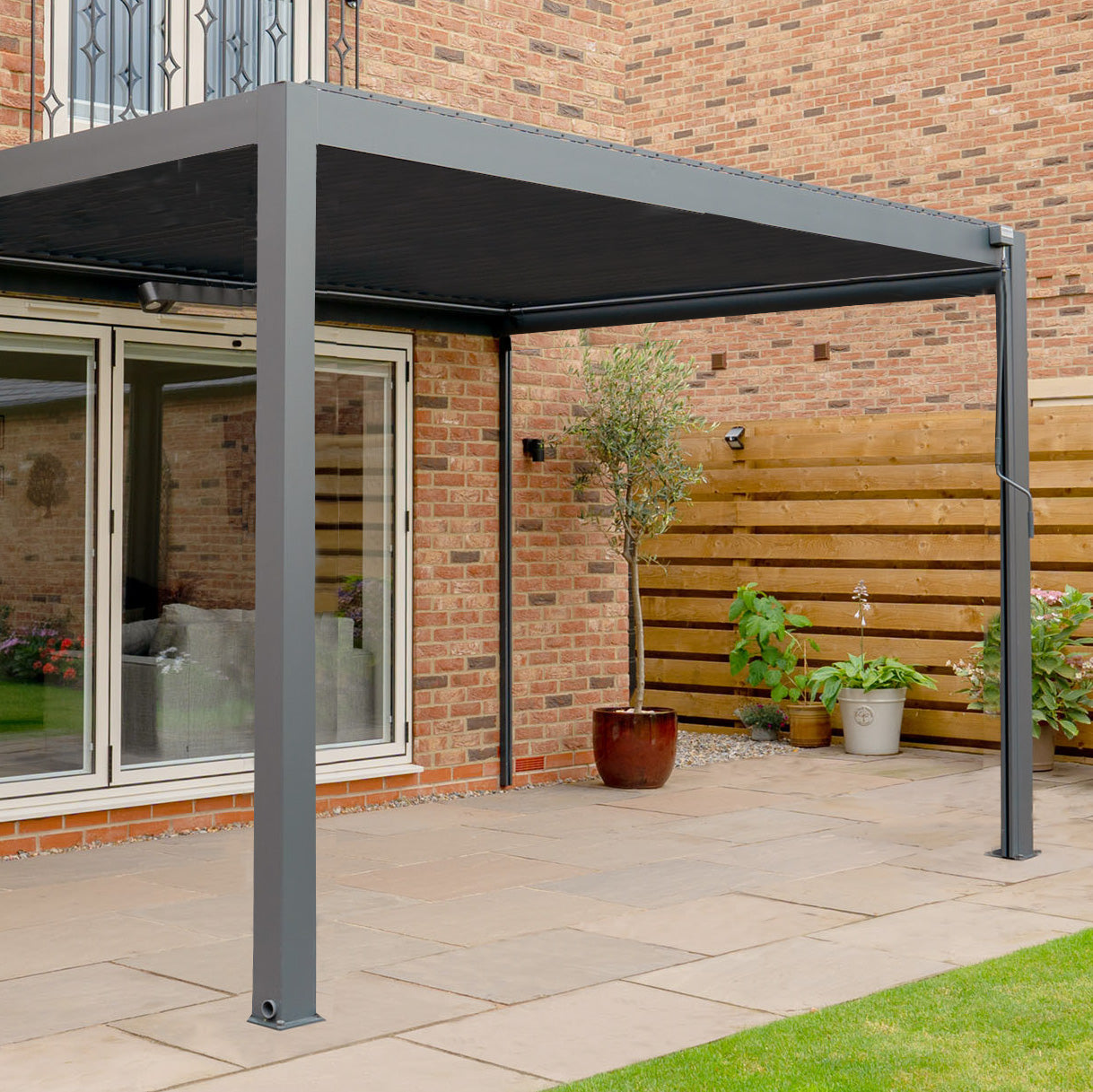 The PergoSTET Plus 3m x 3m grey wall mounted pergola with LED lighting enhances this stone patio beside a brick house and wooden fence, complemented by potted plants.