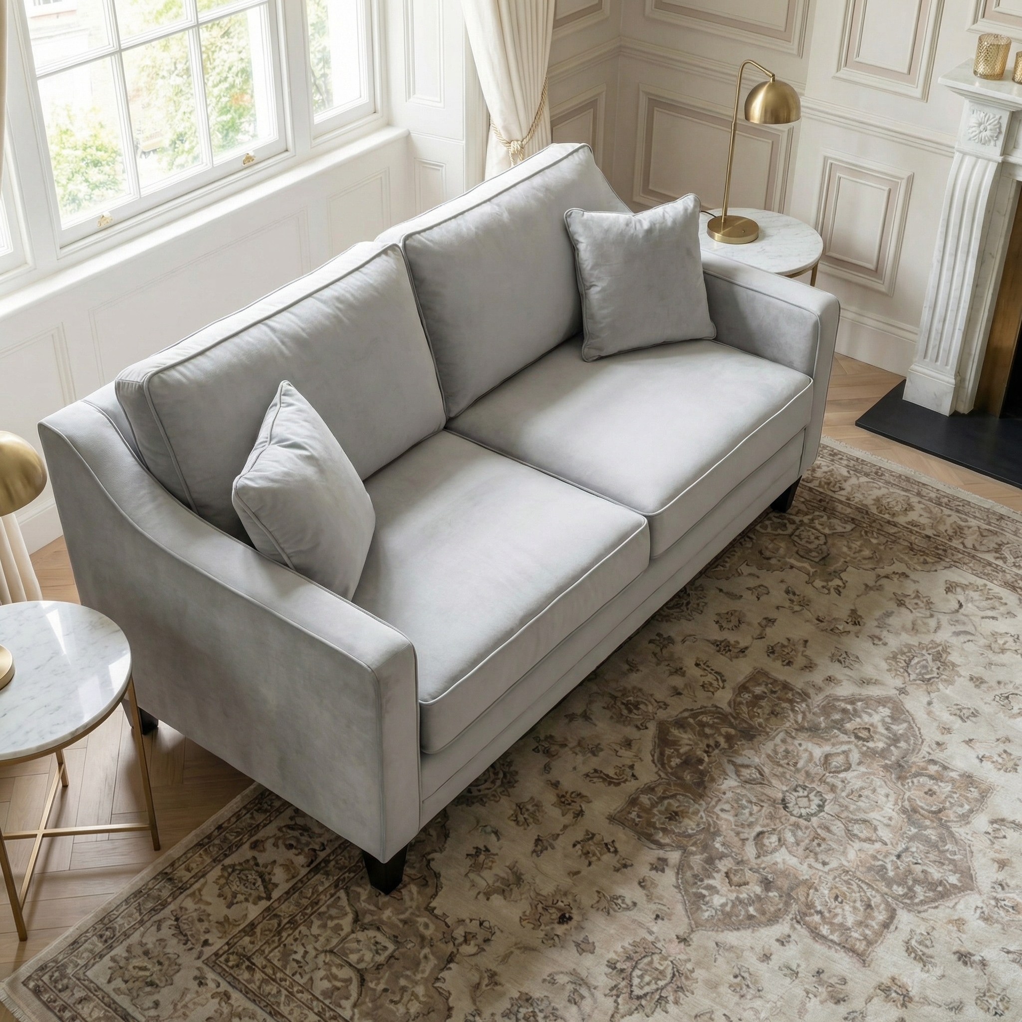 The Wiltshire 2 Seat Velvet Sofa in Feather Grey, paired with two cushions, sits in a bright, elegant living room with a patterned rug and marble side tables.