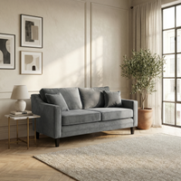 The Wiltshire 2 Seat Velvet Sofa in Steel Grey grounds this airy living room, paired with abstract art, a side table, a potted plant, and a large window.