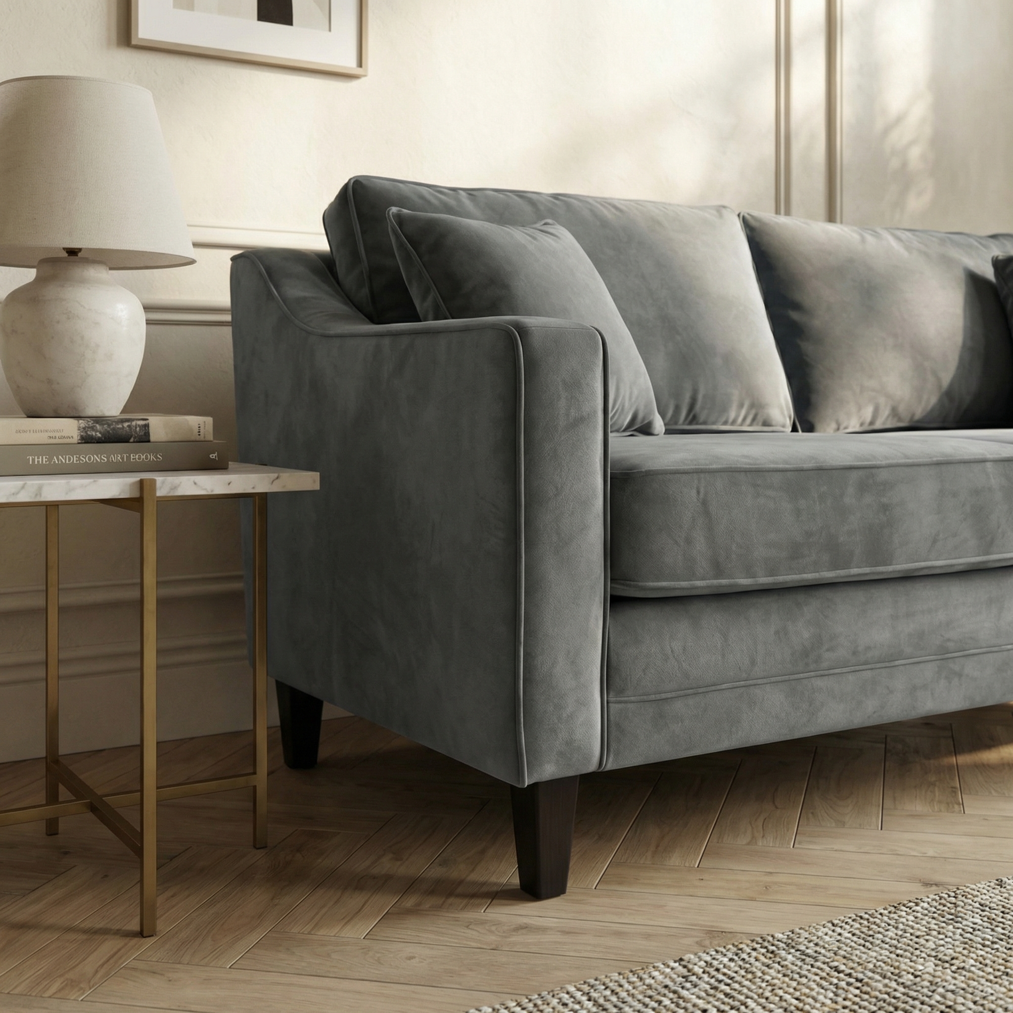 The Wiltshire 2 Seat Velvet Sofa in Steel Grey is placed beside a marble-top side table with a lamp and books, set against a neutral-toned wall.