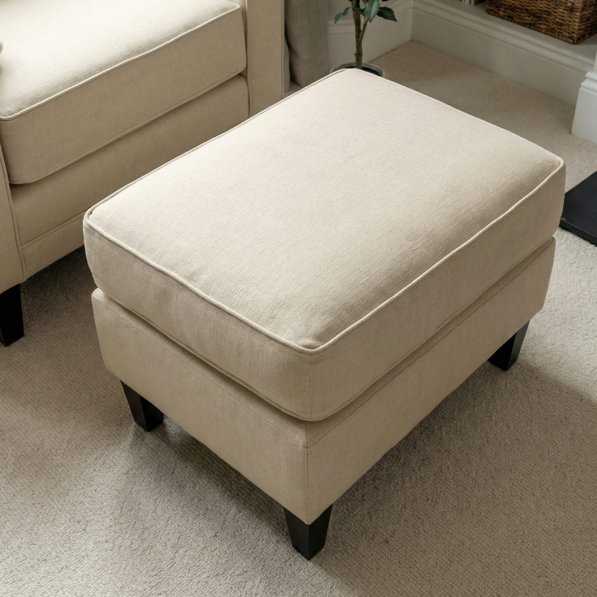 The Wiltshire Footstool in Beige, featuring dark wooden legs, rests elegantly on a light carpeted floor, bringing both comfort and style to your space.