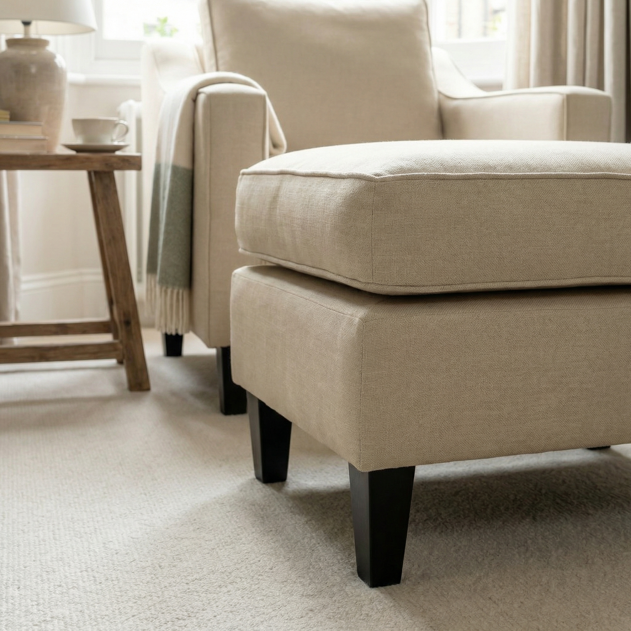 Beige armchair with the Wiltshire Footstool in Beige on a carpeted floor, next to a wooden side table and a window.