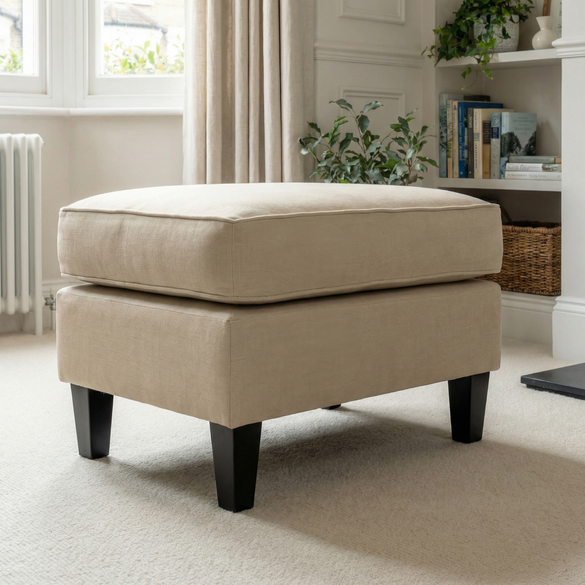 A Wiltshire Footstool in Beige with dark wooden legs is placed in a cozy, carpeted living room near a window and bookshelf.