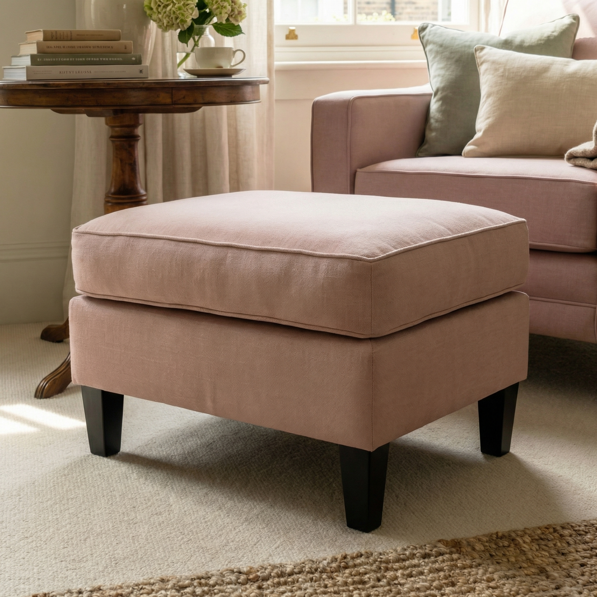 The Wiltshire Footstool in Dusky Pink with dark wooden legs sits in a cozy living room near a sofa, table, and window.