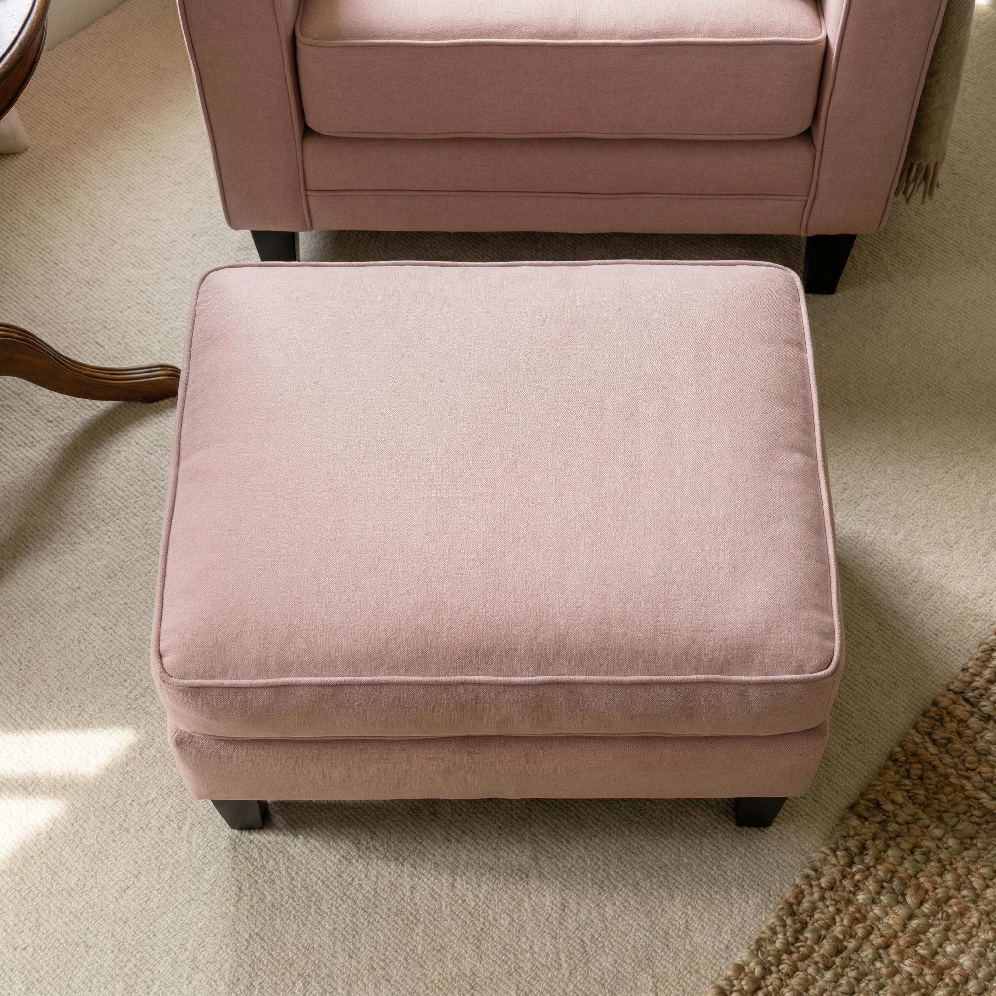 The Wiltshire Footstool in Dusky Pink, upholstered in soft fabric, sits on a light carpet near a matching armchair and wooden table—ideal as a handcrafted accent for any cozy space.