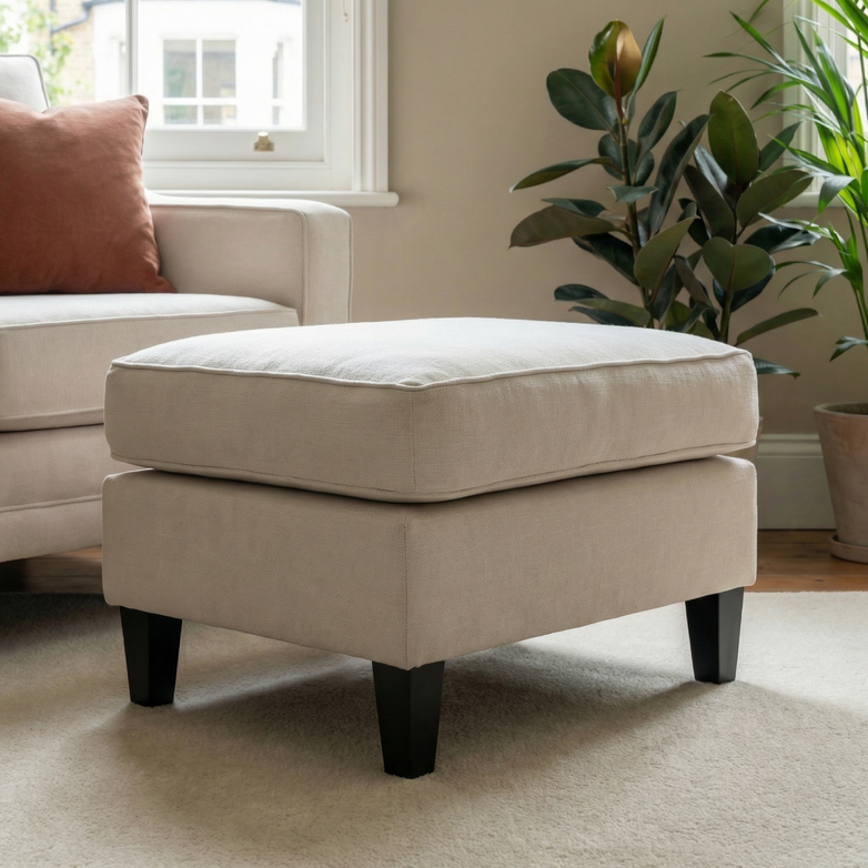 The Wiltshire Footstool in Ivory with dark legs adds comfort and style to a cozy living room, placed near a sofa, lush plants, and a window for an elegant touch.