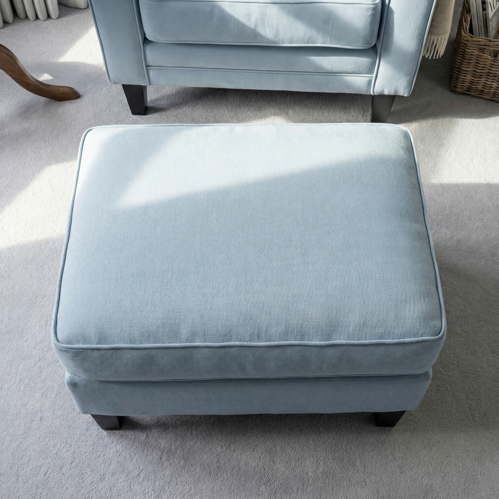 Wiltshire Footstool in Sky Blue placed on a beige carpet with sunlight streaming in, featuring part of the matching chair in the background.
