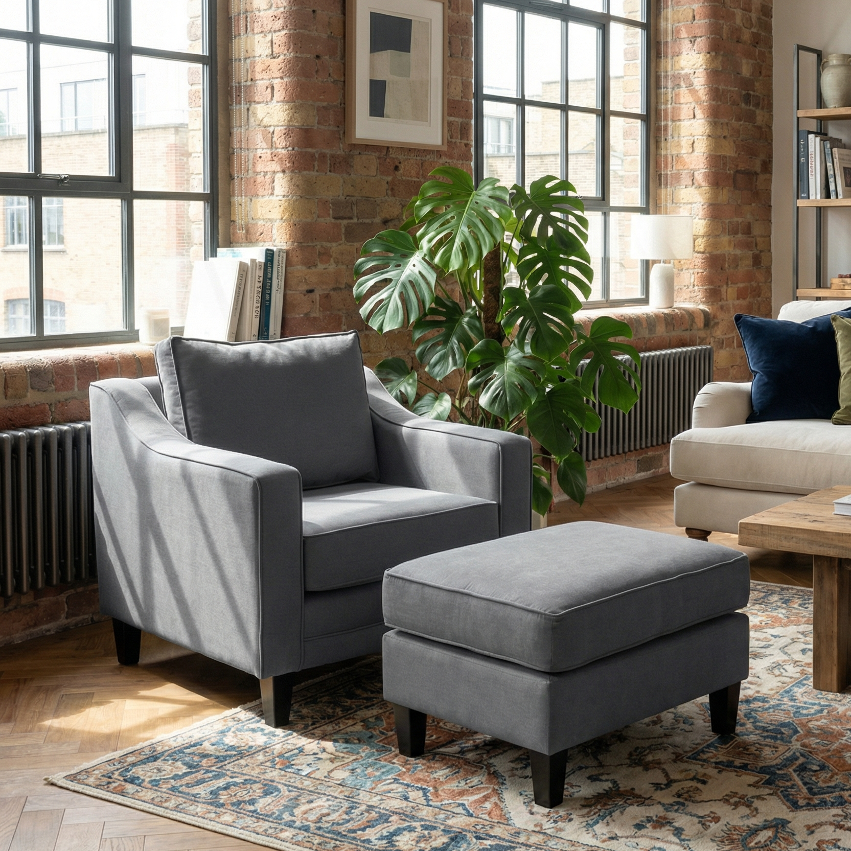Gray armchair with a Wiltshire Footstool in Smoke Grey, set in a cozy living room with brick walls, large windows, and a leafy plant.