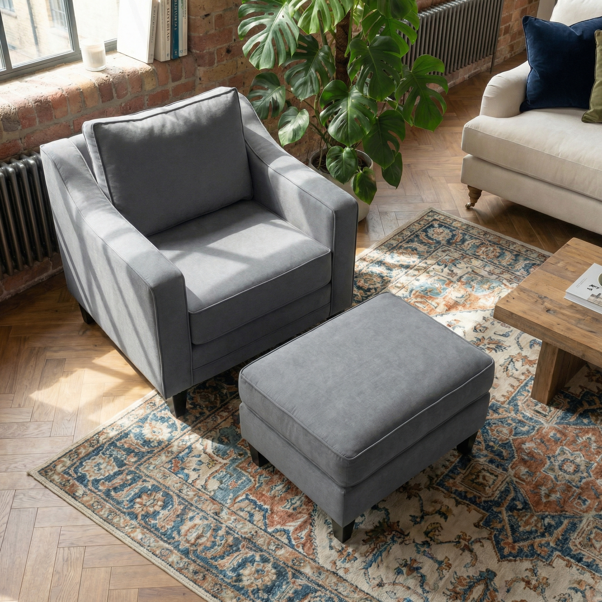 Gray armchair with a Wiltshire Footstool in Smoke Grey on a patterned rug, beside a wooden coffee table and a green plant.