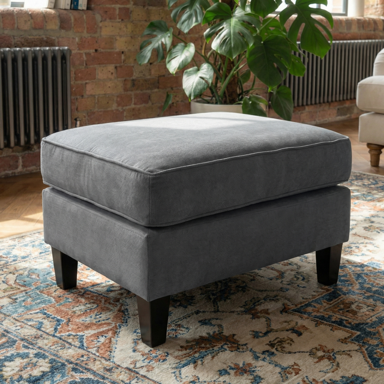 The Wiltshire Footstool in Smoke Grey, featuring dark wooden legs, sits on a patterned rug in a cozy, sunlit living room, bringing modern style to the space.