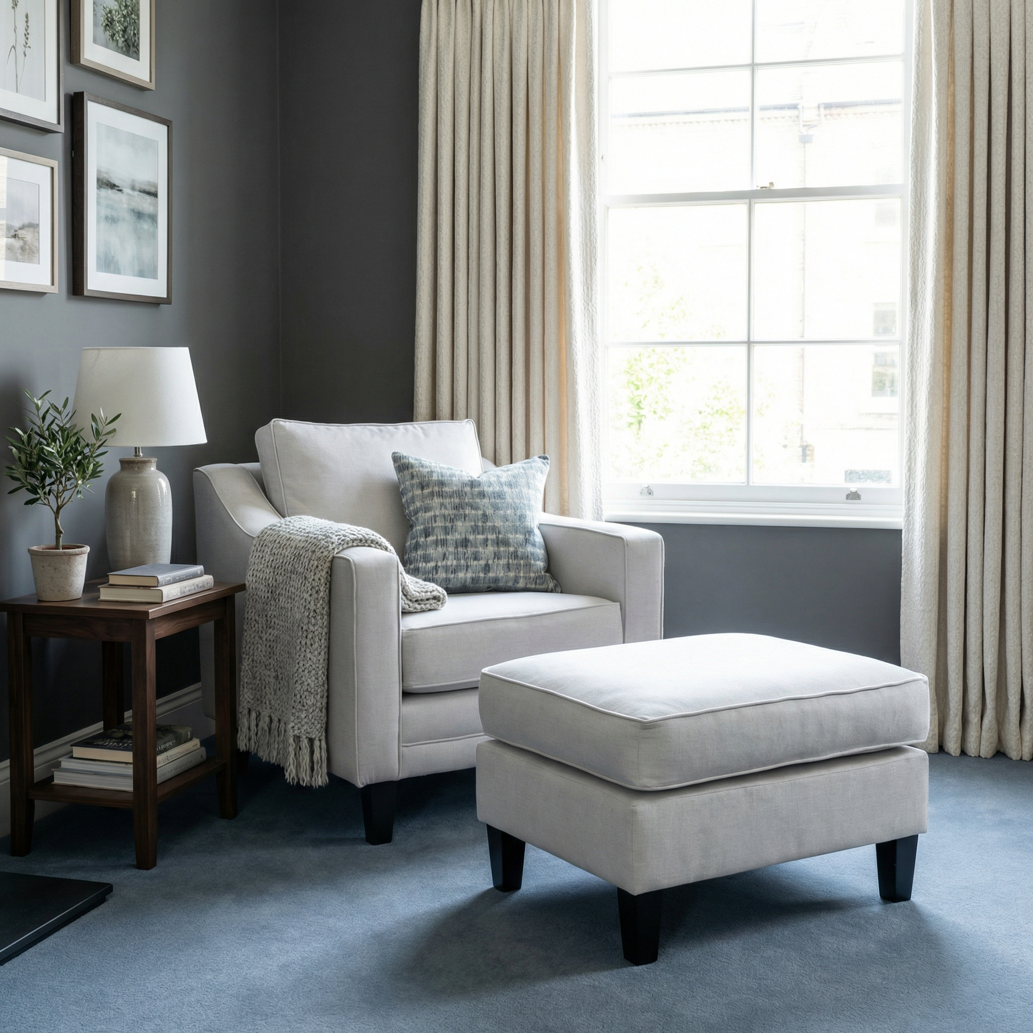 A cozy living room with a beige armchair, Wiltshire Footstool in Warm Grey, side table, lamp, and a large window with curtains.
