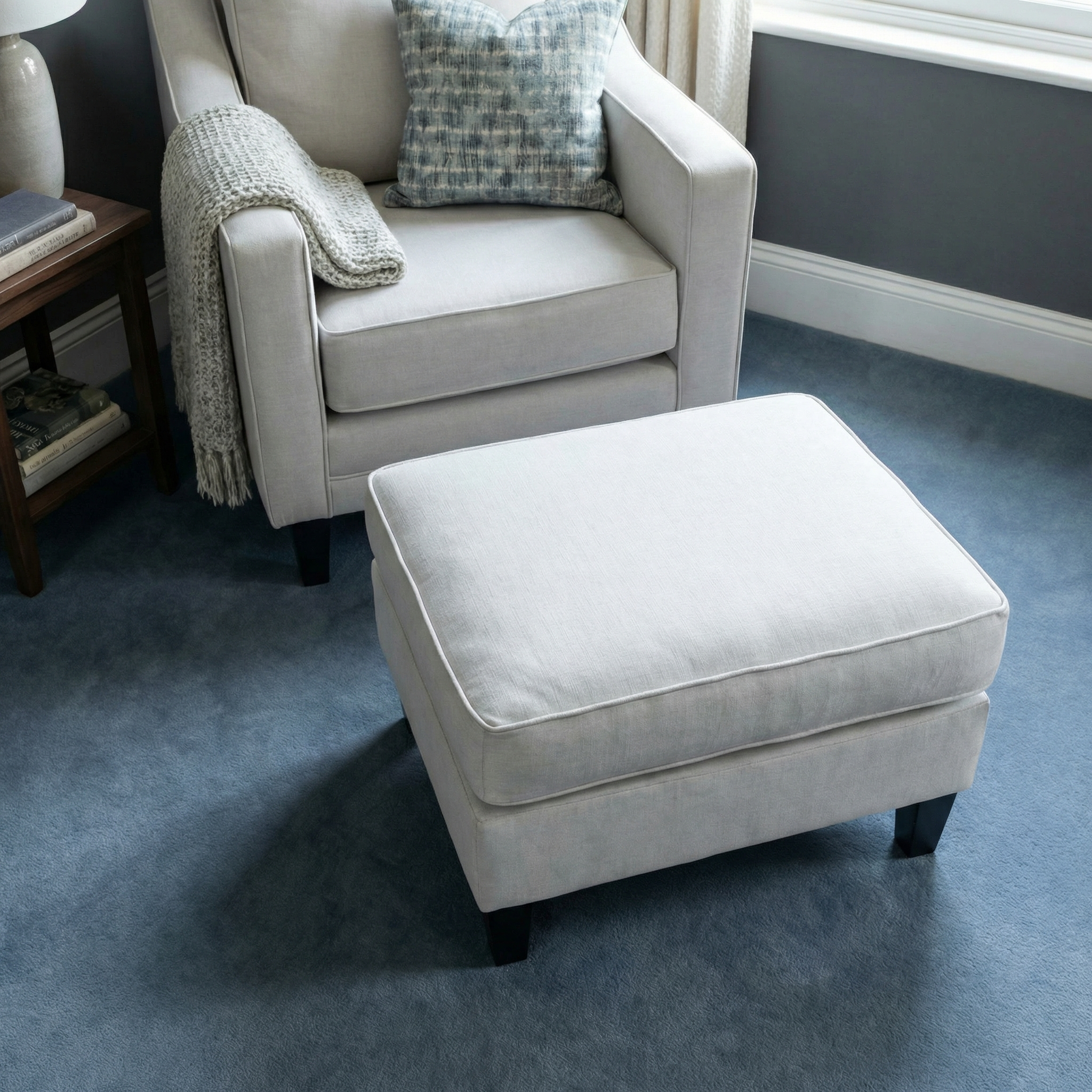 Light gray armchair with a Wiltshire Footstool in Warm Grey sits on blue carpet, topped by a cozy throw blanket and cushion.