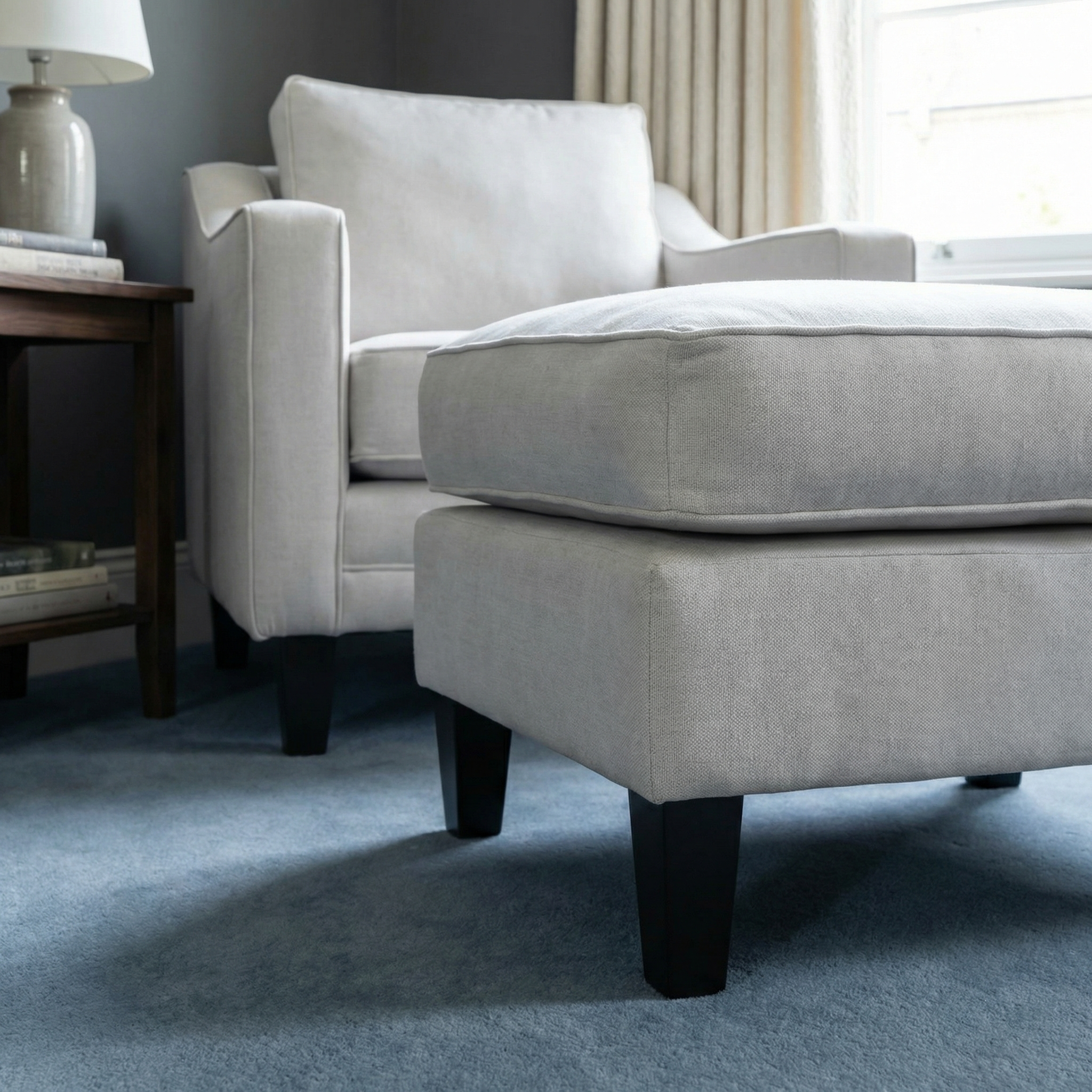 Light gray armchair and Wiltshire Footstool in Warm Grey with dark legs on a blue carpet in a cozy, softly lit room.