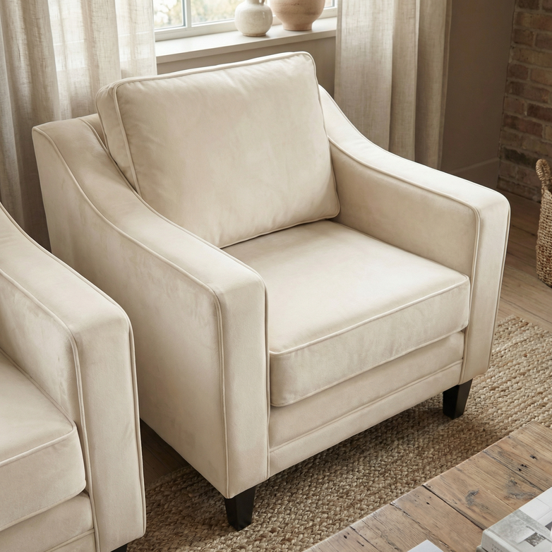 The Wiltshire Velvet Armchair in Cream, featuring a plush cushion and sustainable wood frame, handmade in England, sits on a woven rug in a cozy, sunlit living room.