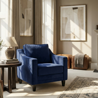 The Wiltshire Velvet Armchair in Royal Blue sits in a sunlit living room with neutral decor, abstract art, and a side table topped with a lamp.