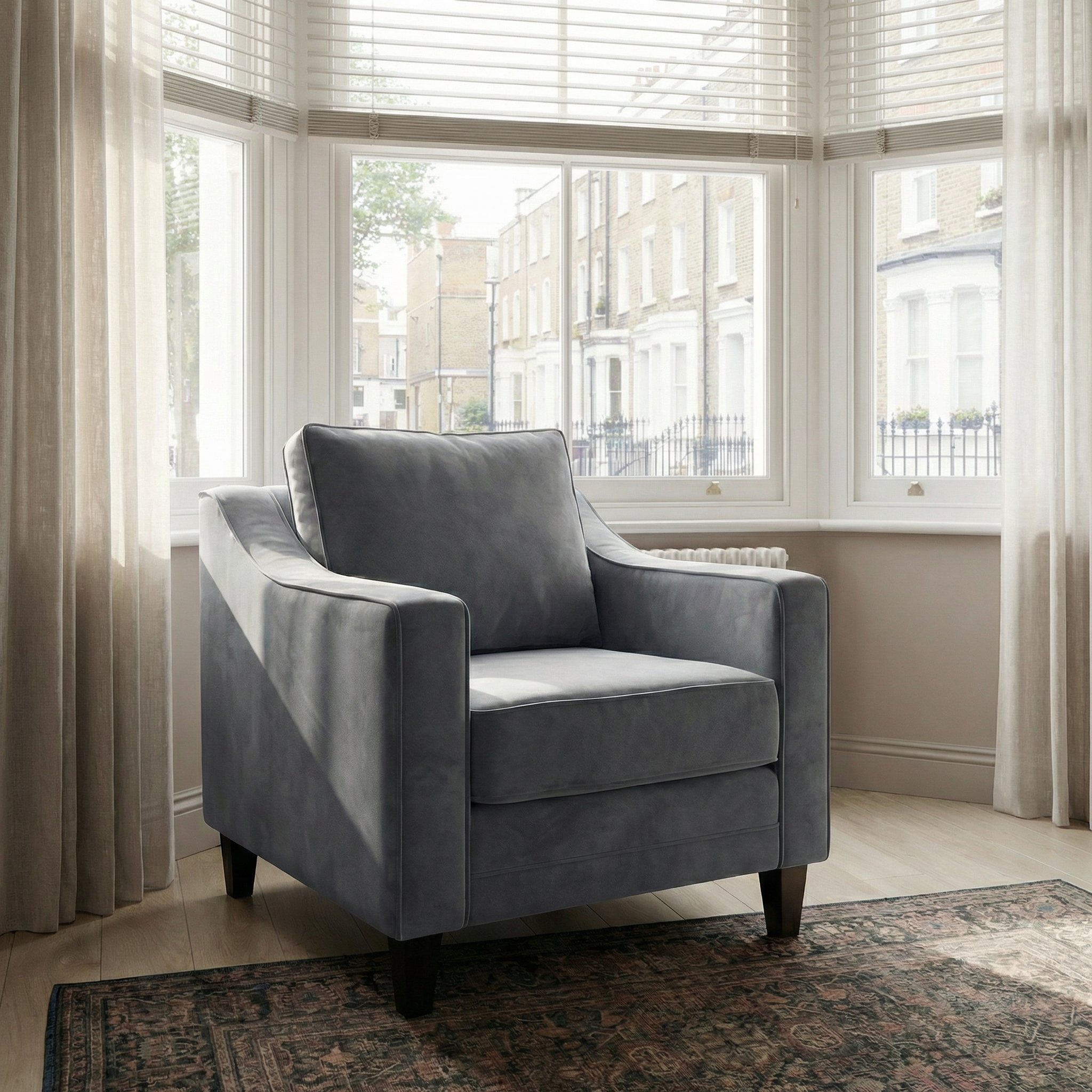 A Wiltshire Velvet Armchair in Steel Grey sits by a bay window with sheer curtains in a bright, cozy room.