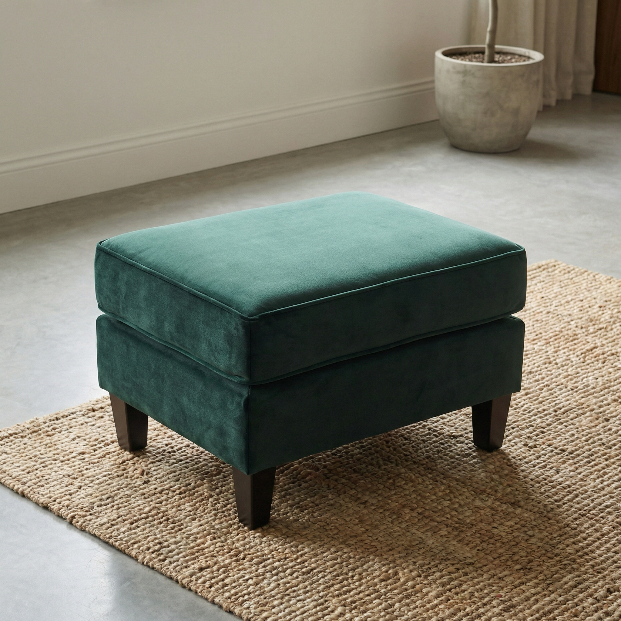 The Wiltshire Velvet Footstool in Bottle Green with dark wooden legs sits on a woven rug in a modern, minimal room, complemented by a plant pot.