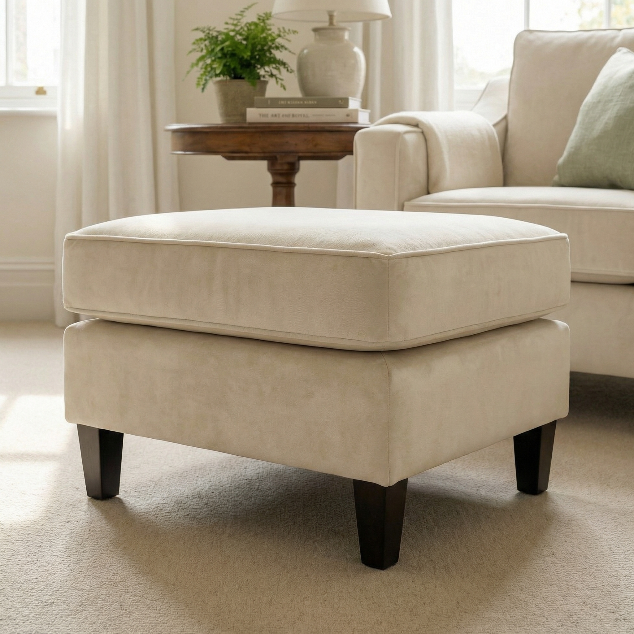 The Wiltshire Velvet Footstool in Cream, featuring dark wooden legs, brings comfort and style to a bright, cozy living room.