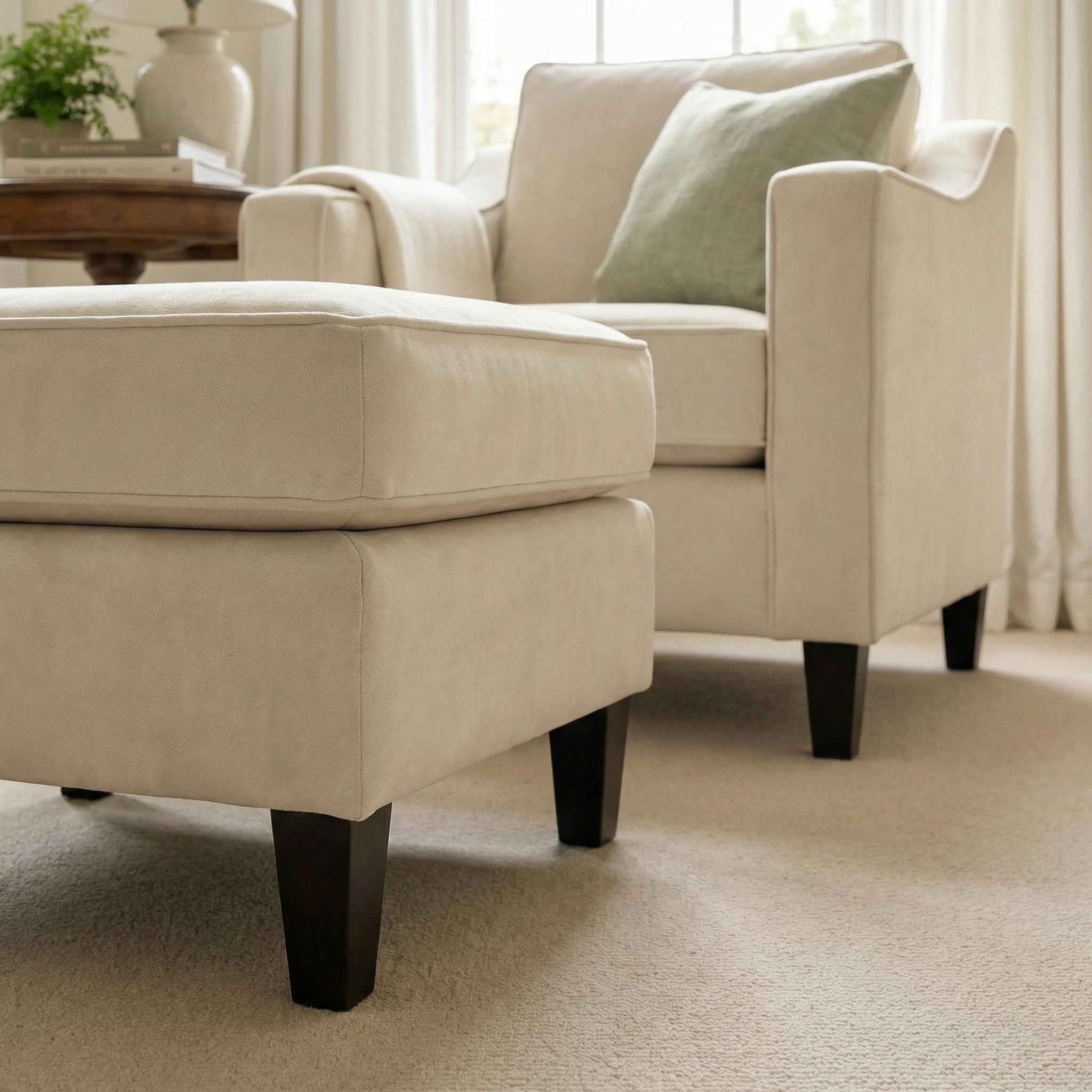 Cream-colored armchair and ottoman with dark legs, paired with the Wiltshire Velvet Footstool in Cream, in a bright living room with soft light and neutral decor.