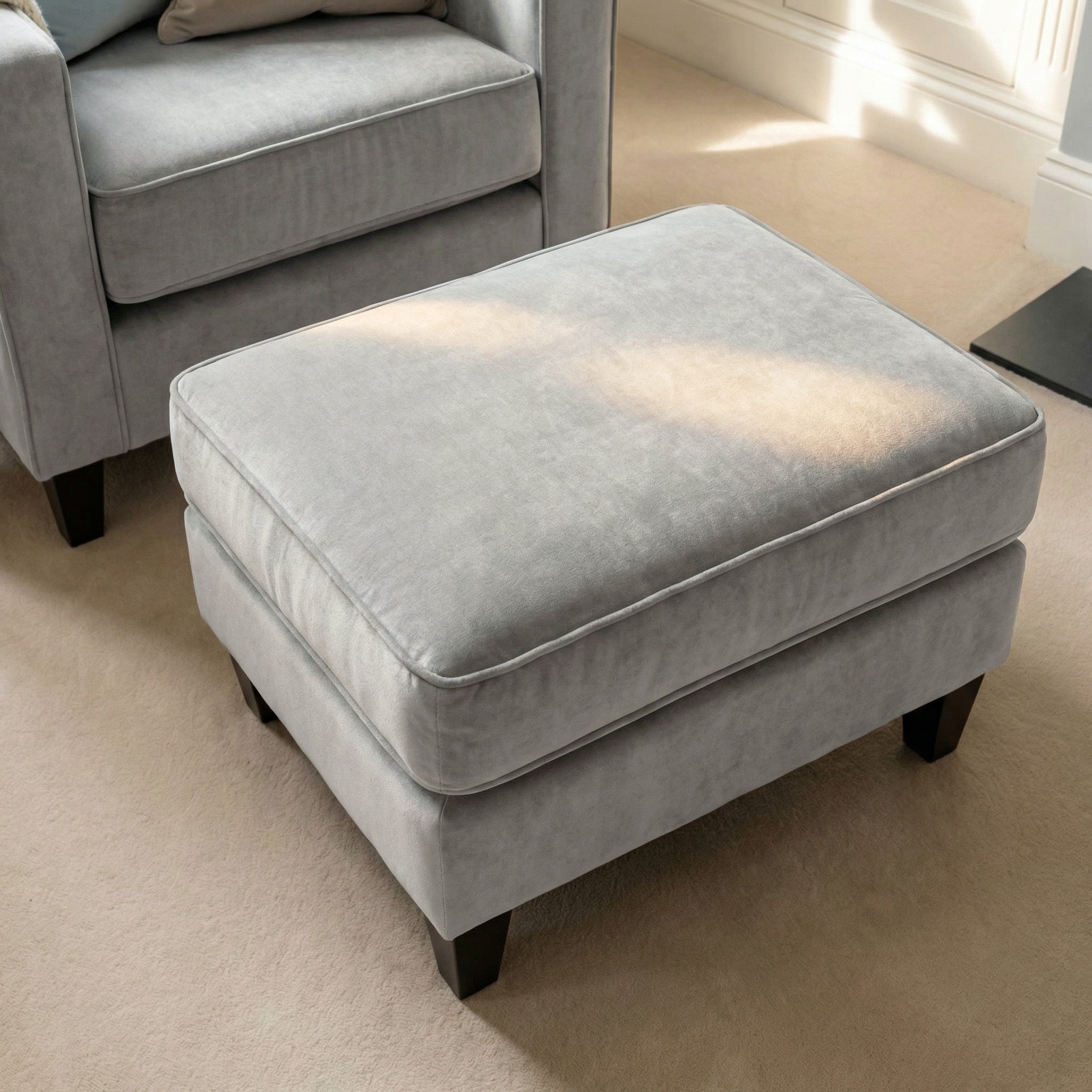 The Wiltshire Velvet Footstool in Feather Grey with dark wooden legs rests on a beige carpet beside a matching armchair, adding comfort and style to the space.