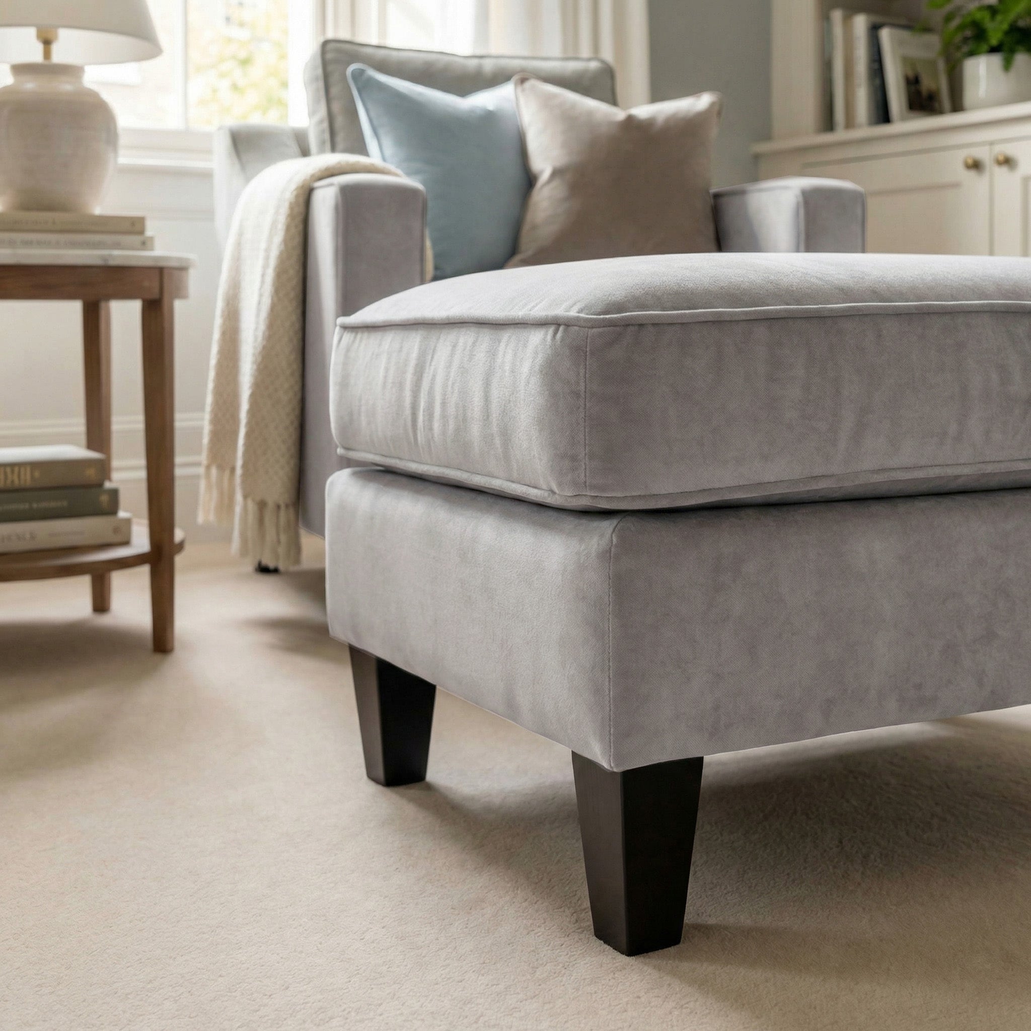 Light gray upholstered armchair with the Wiltshire Velvet Footstool in Feather Grey in a cozy, sunlit living room featuring neutral decor.
