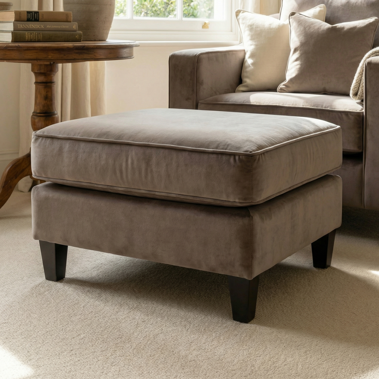 A Wiltshire Velvet Footstool in Mink with dark legs sits on a beige carpet in a cozy, sunlit living room.