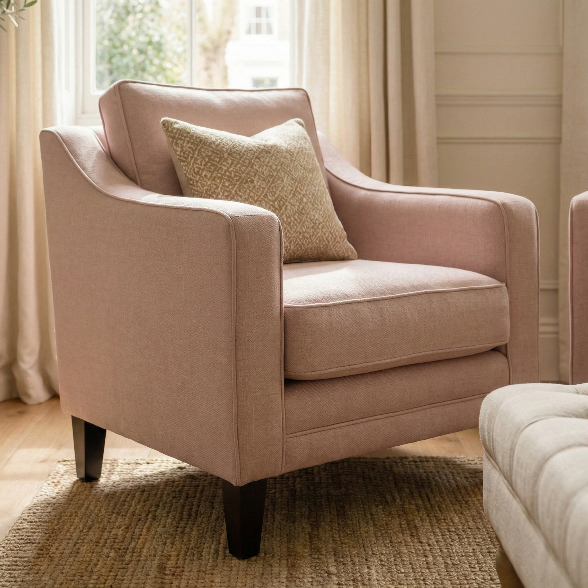 The Wiltshire Armchair in Dusky Pink, adorned with a patterned throw pillow, sits on a woven rug in a sunlit room.