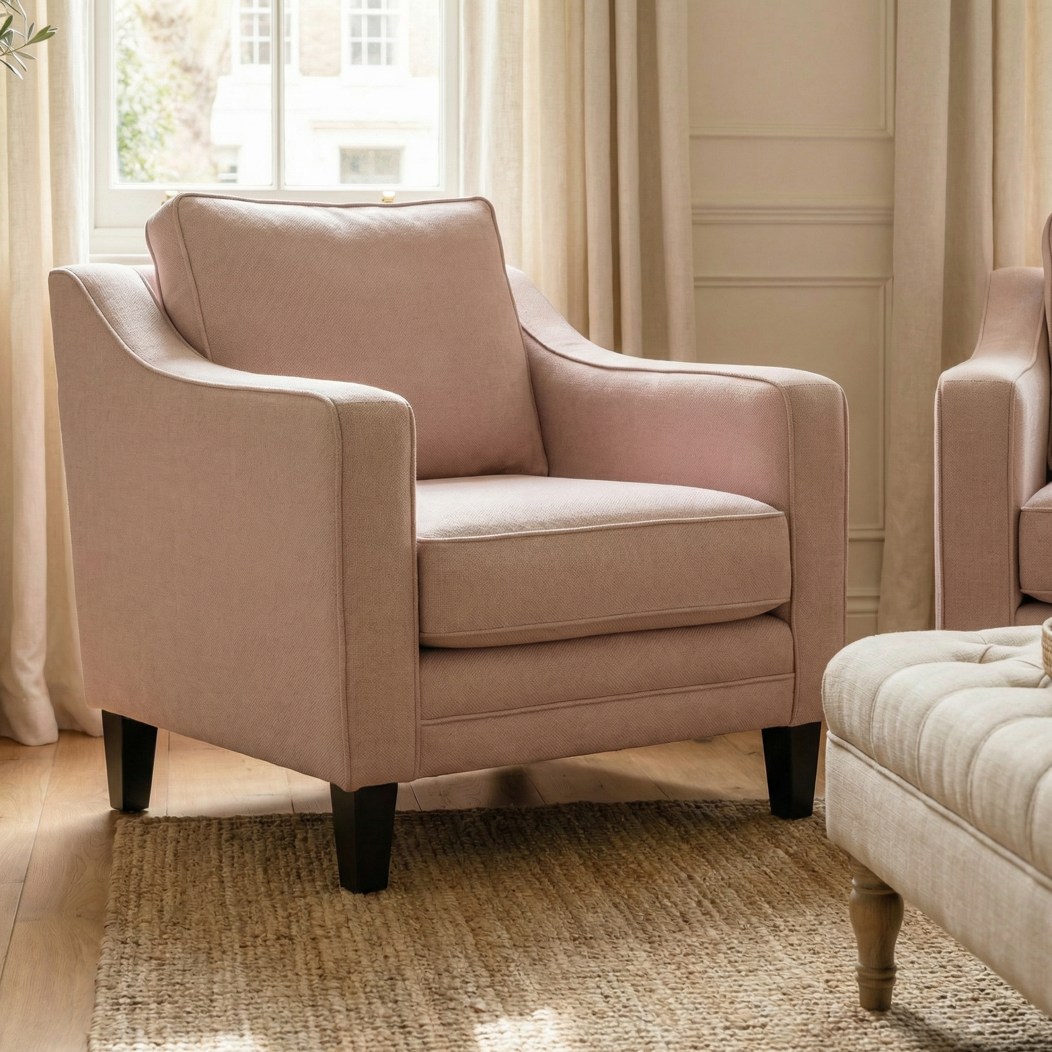 The Wiltshire Armchair in Dusky Pink with dark wooden legs is placed on a woven rug, adding charm to a bright and cozy living room.