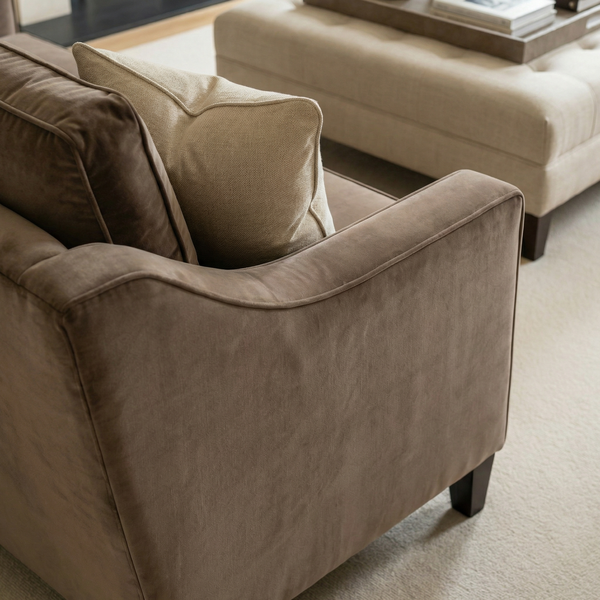 Brown suede couch with a beige pillow beside a cream ottoman on a light carpeted floor, complemented by the Wiltshire Velvet Armchair in Mink for added elegance.