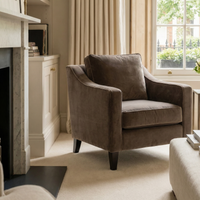A Wiltshire Velvet Armchair in Mink is placed in a cozy, sunlit living room beside a window with cream curtains.