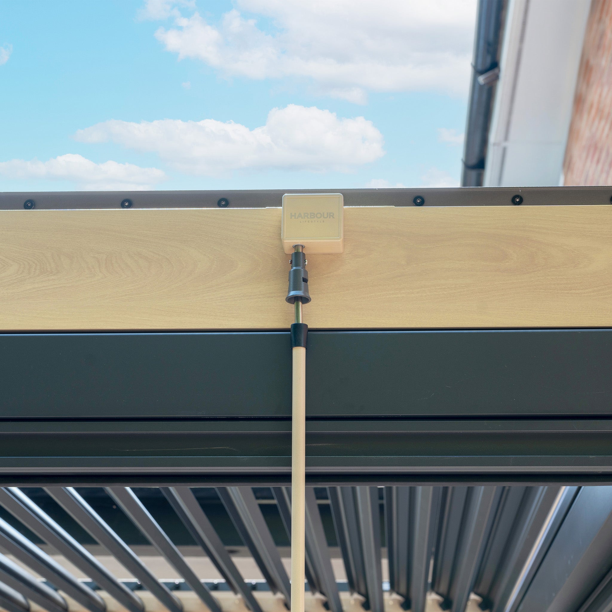 Close-up of the PergoSTET Plus 3m x 3m Square Pergola in Wood Effect, showing tilting louvres, manual blinds, LED lighting, and a beige control box with cable underneath.