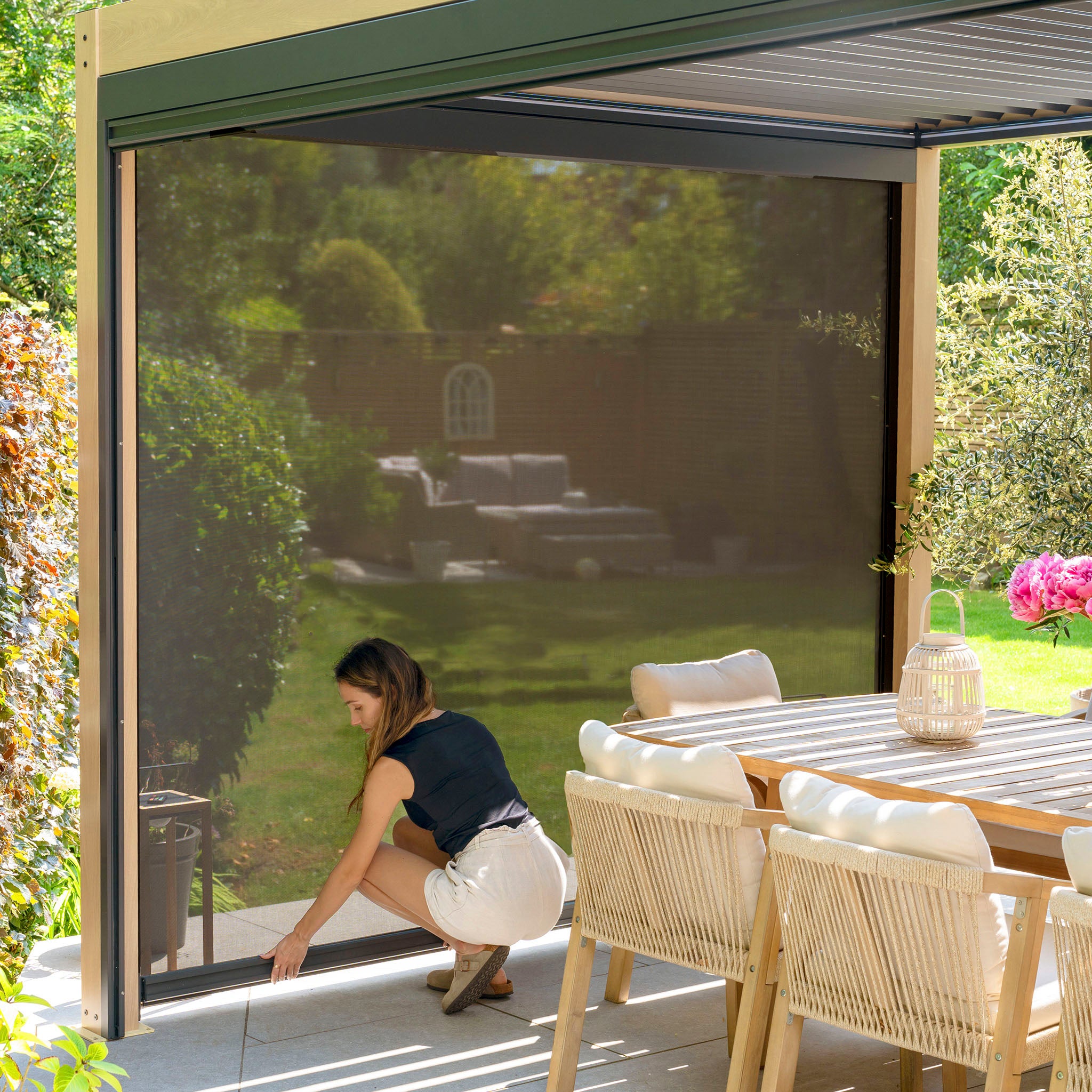 A woman adjusts the screen beneath a modern PergoSTET Plus pergola, featuring adjustable louvres and LED lighting, in a sunny patio. The setup, including table and chairs, overlooks a lush garden for ideal comfort and style.