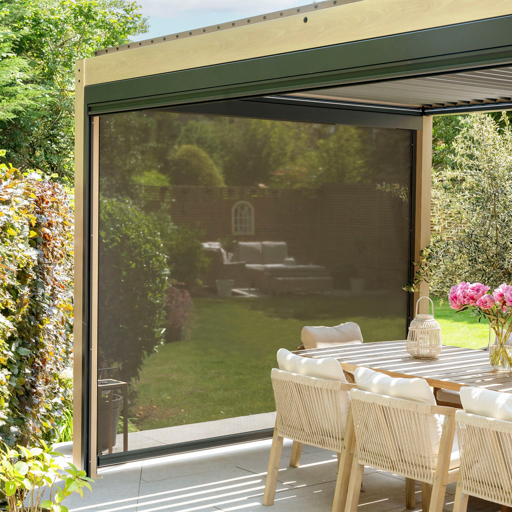 An outdoor dining area with a wooden table and wicker chairs is enhanced by the PergoSTET Plus 3m x 4m Rectangular Pergola, featuring adjustable louvres, three drop sides, LED lighting with wood effect, potted flowers, lush greenery, and a retractable screen for ambiance and flexible shade.