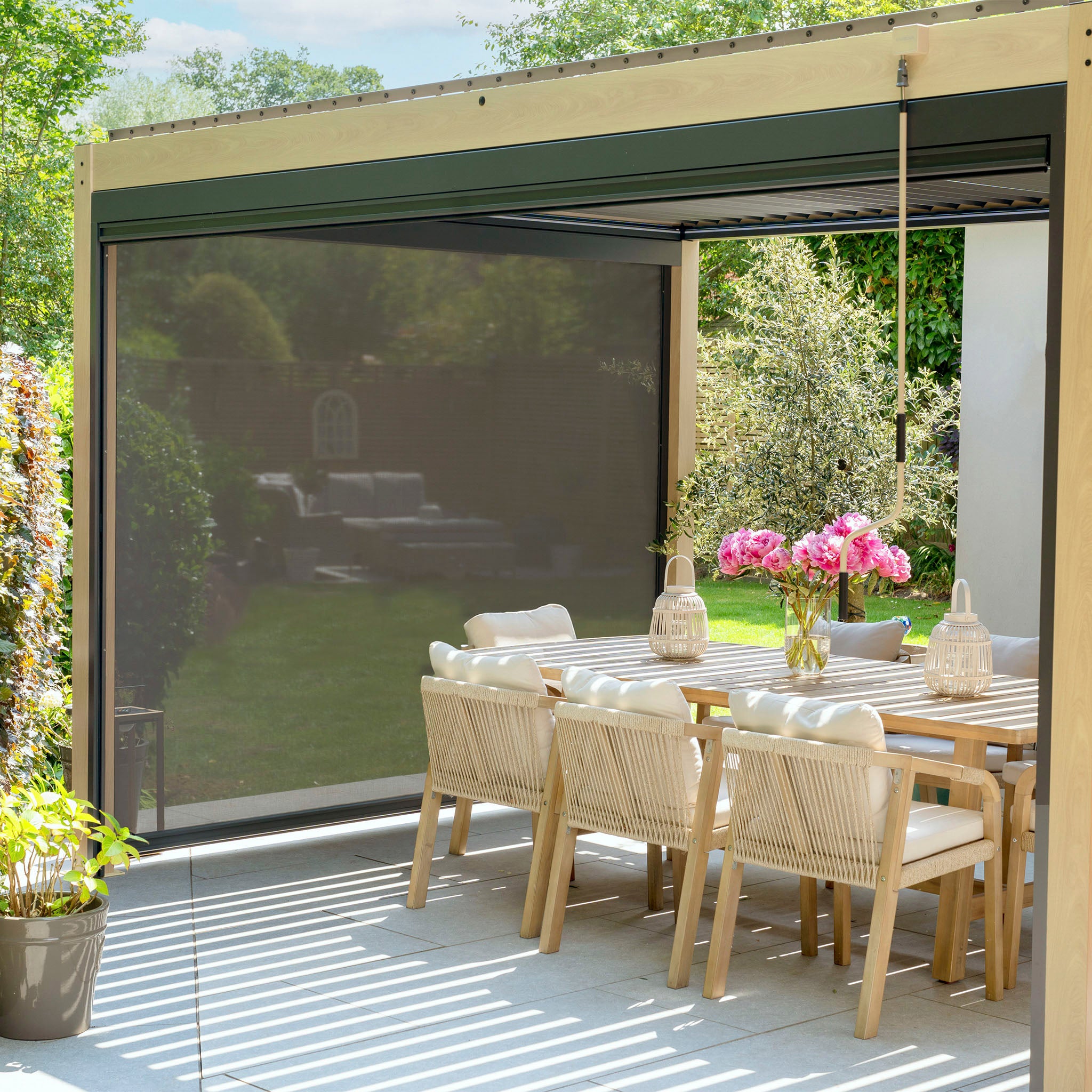The outdoor dining area features a wooden table and chairs with a pink flower centerpiece, enclosed by the PergoSTET Plus 3m x 4m Pergola with adjustable louvres and LED lighting, offering a sunny garden view.