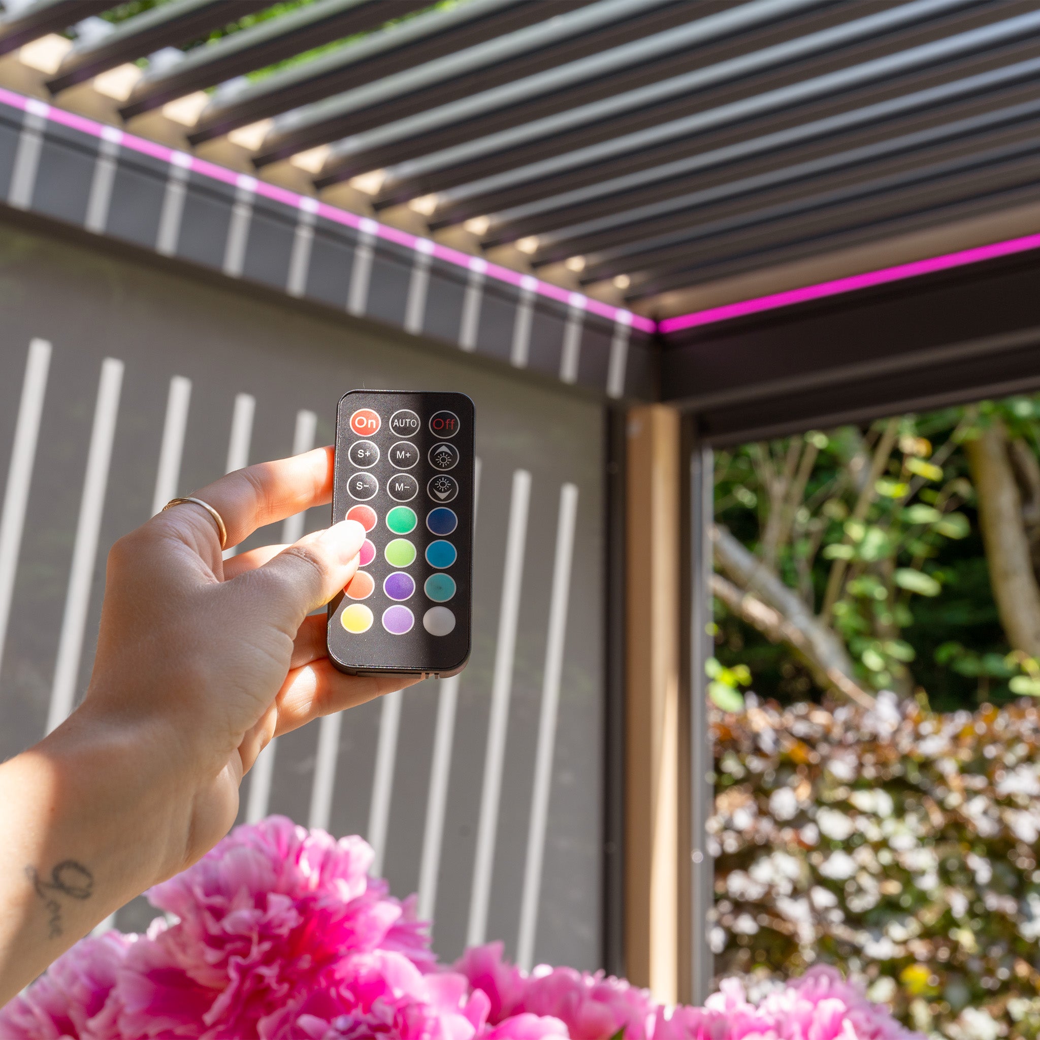 A hand holds a remote control in an elegant outdoor area with pink flowers and a PergoSTET Plus pergola, featuring adjustable louvres and LED lighting that combine style and functionality.