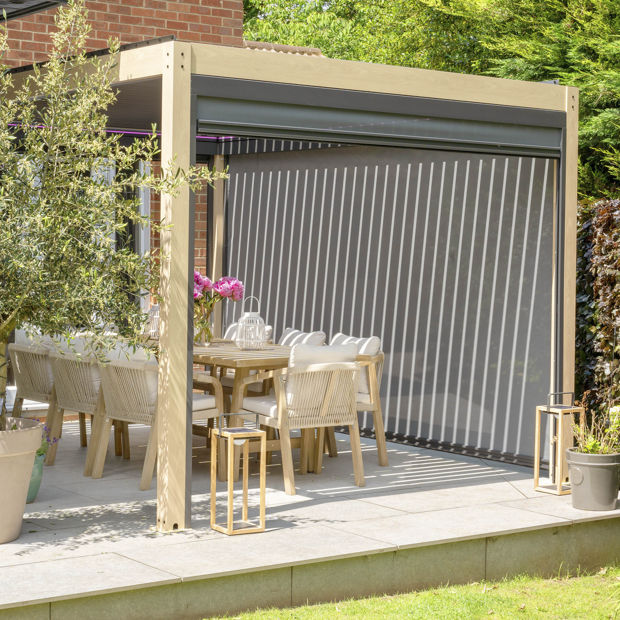 Outdoor patio showcasing a PergoSTET Plus 3m x 4m rectangular pergola with wood effect, adjustable louvres, and LED lighting, complemented by a dining set and potted plants under the sun.