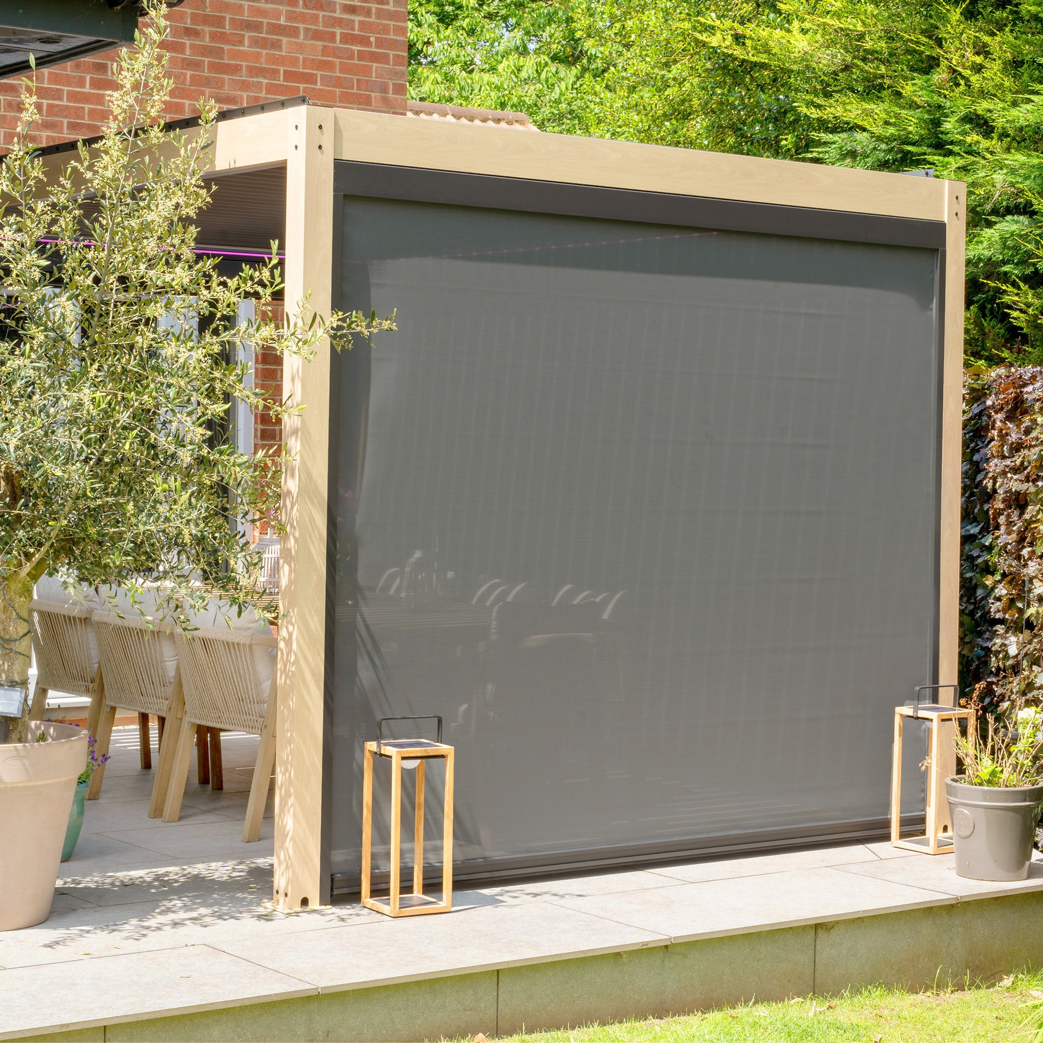 Outdoor patio with a PergoSTET Plus 3m x 4m Rectangular Pergola featuring 3 drop sides, LED lighting, potted plants, and wooden chairs around a dining table.