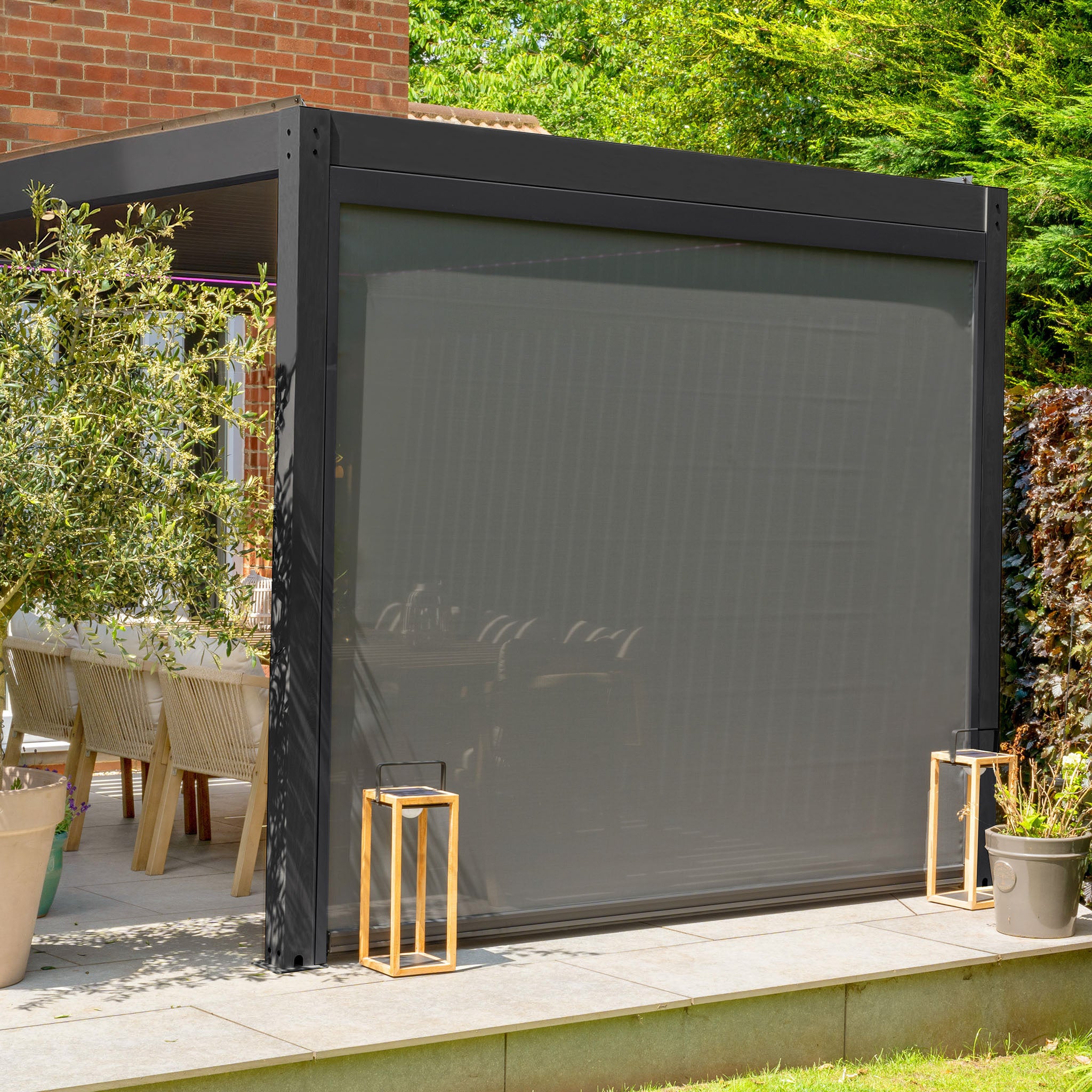 A modern patio featuring the PergoSTET Plus 3m x 3m Square Pergola in Grey, complete with adjustable waterproof roof, retractable screen, dining set, lush plants, ambient lanterns on a stone deck, and LED lighting for year-round enjoyment and sophistication.