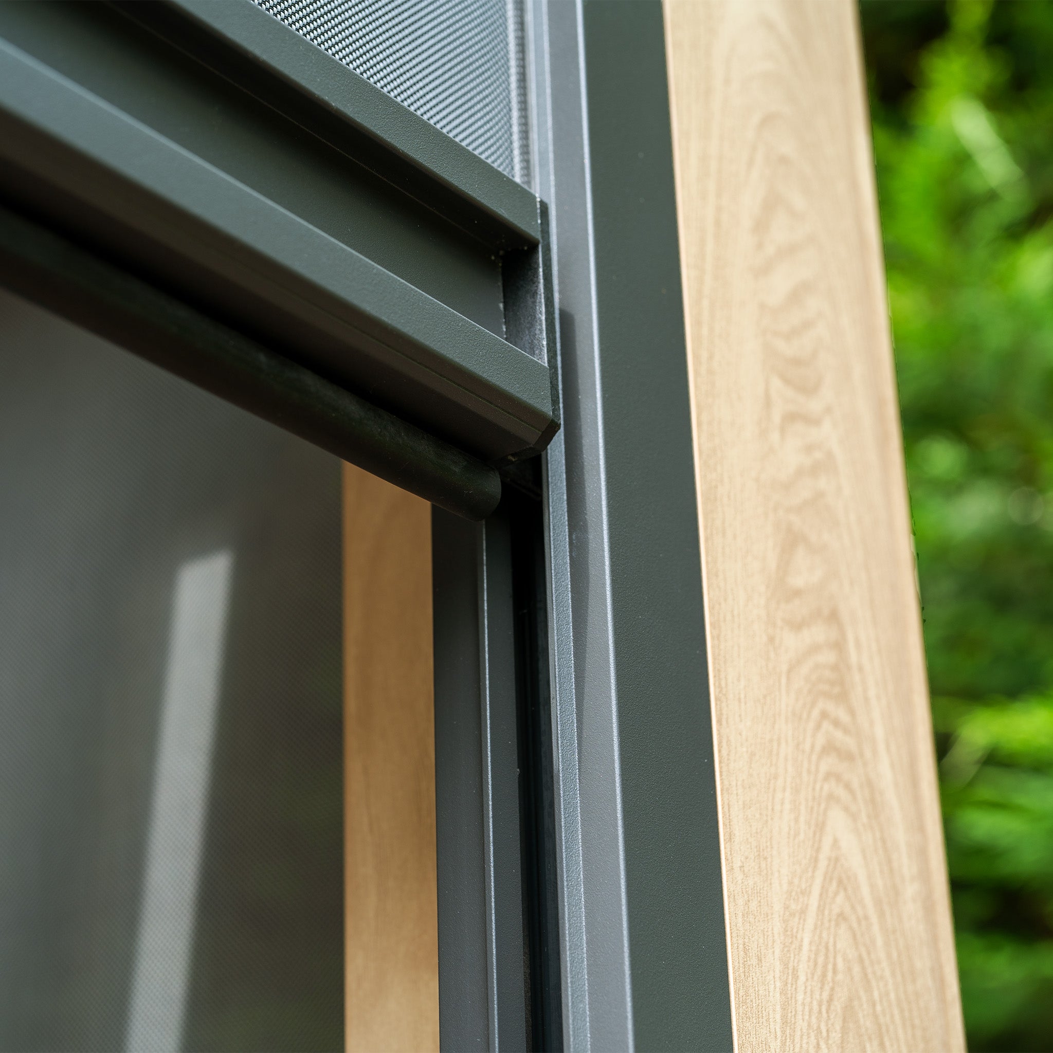 Close-up of a modern window frame with wood and metal detailing, reminiscent of the PergoSTET Plus 3m x 4m Rectangular Pergola with wood effect, set against a blurred green background.