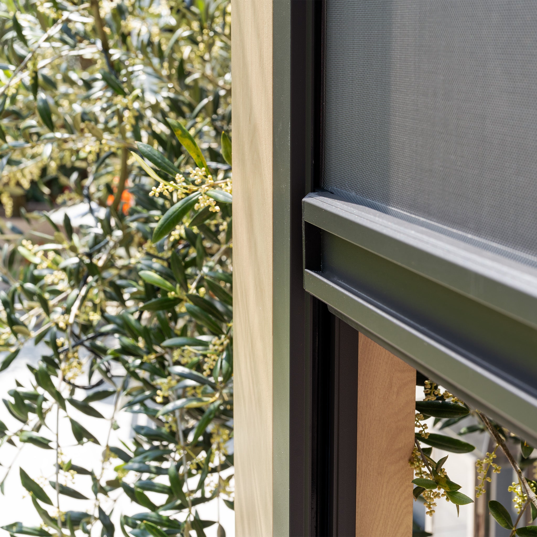 A close-up showcases a corner of the PergoSTET Plus 3m x 4m Rectangular Pergola with its wood-effect frame, adjacent to olive tree branches. Adjustable louvres filter the light, adding elegance to this harmonious scene, enhanced by LED lighting and three drop sides.