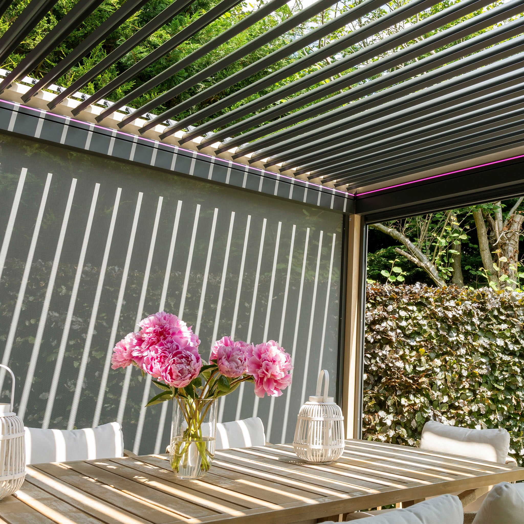 The PergoSTET Plus 3m x 4m Rectangular Pergola, featuring adjustable louvres, wood effect, LED lighting, and three drop sides, casts striped shadows on a table adorned with pink flowers; lanterns sit beside it.