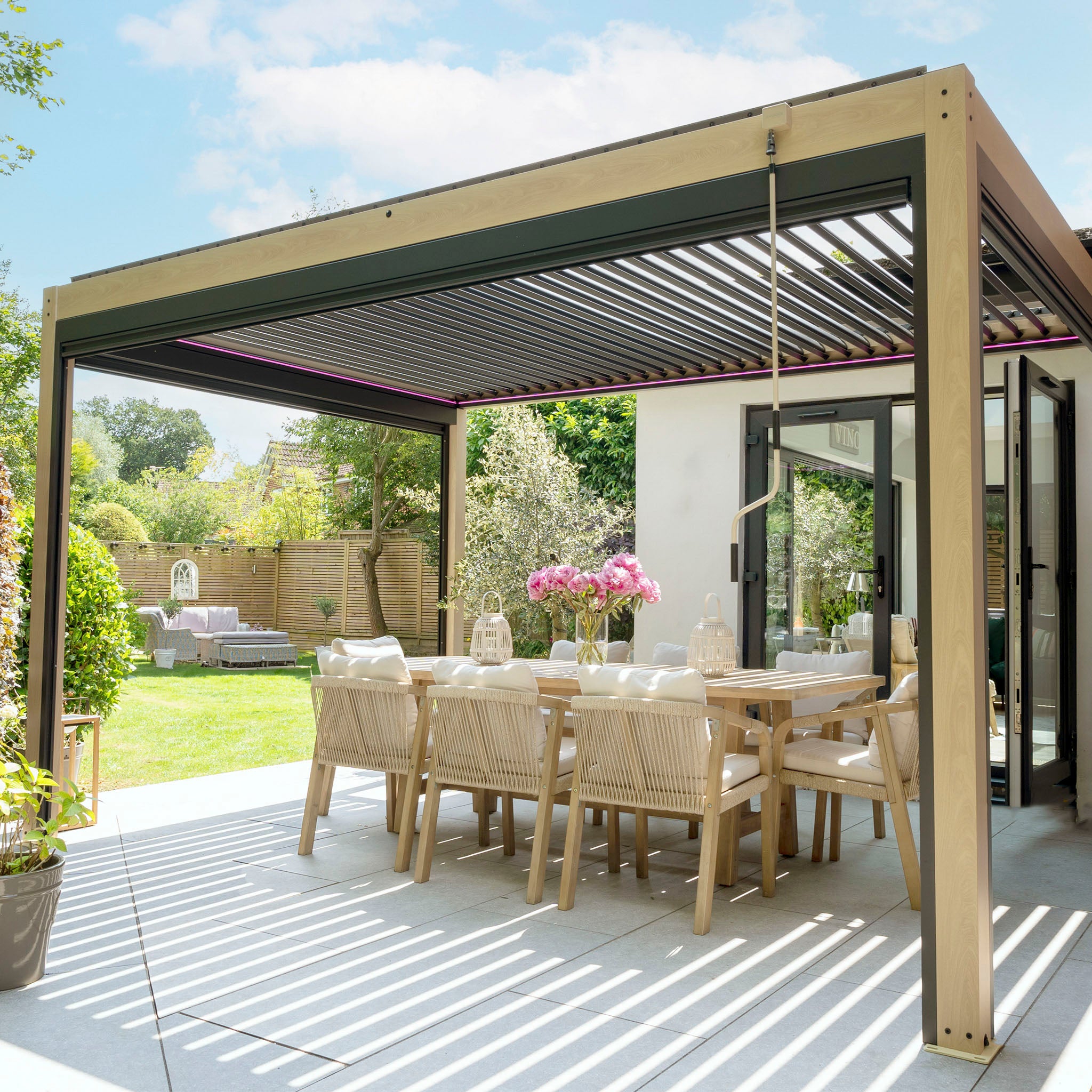 A modern outdoor dining area showcases a sleek PergoSTET Plus 3m x 4m rectangular pergola with adjustable louvres, 3 drop sides, and LED lighting in a wood effect, complemented by a wooden table and chairs amidst a lush garden on a sunny day.