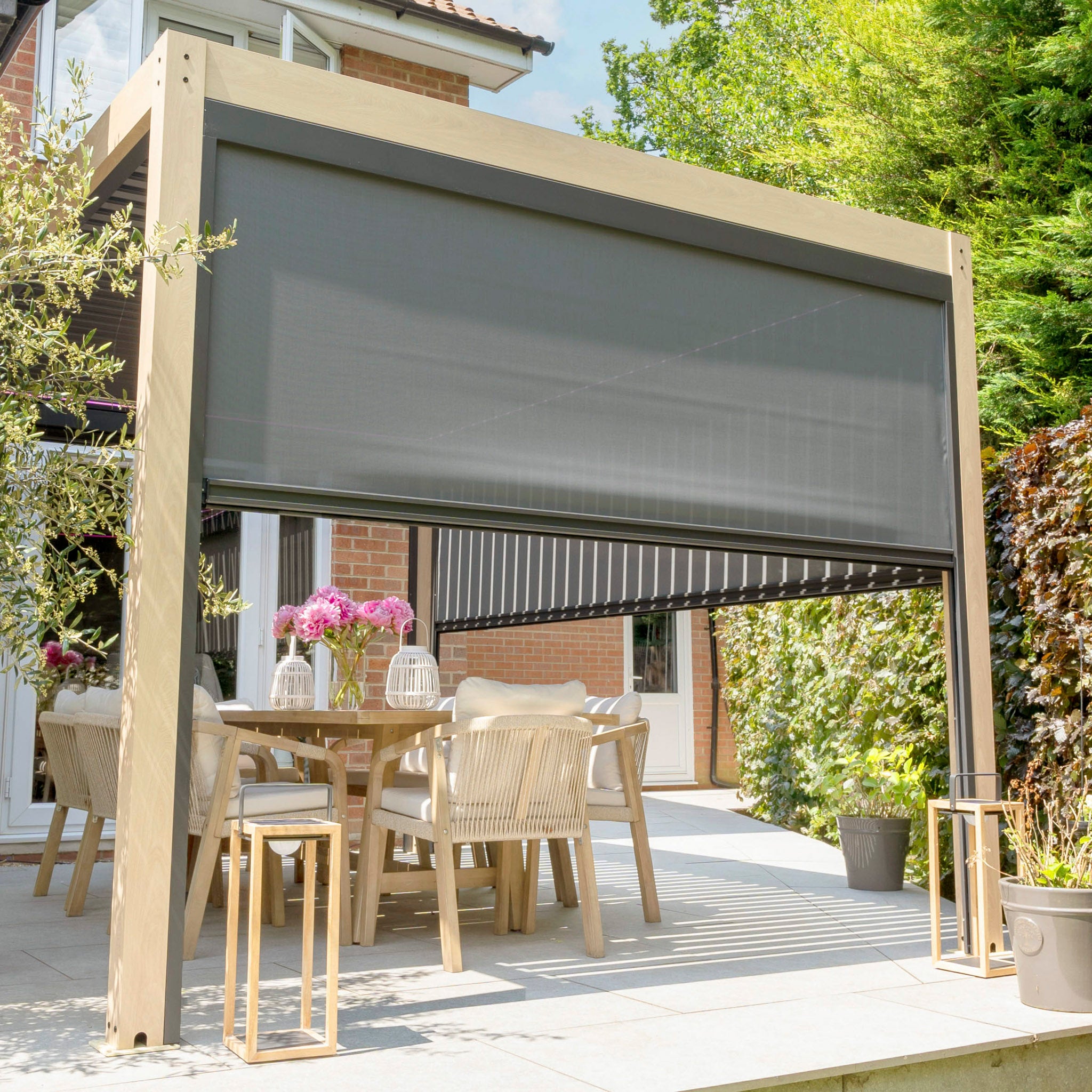 A modern patio showcases a PergoSTET Plus 3m x 4m pergola with 3 drop sides and LED lighting, enhanced by a dining table, chairs, vibrant pink flowers, and lush greenery.