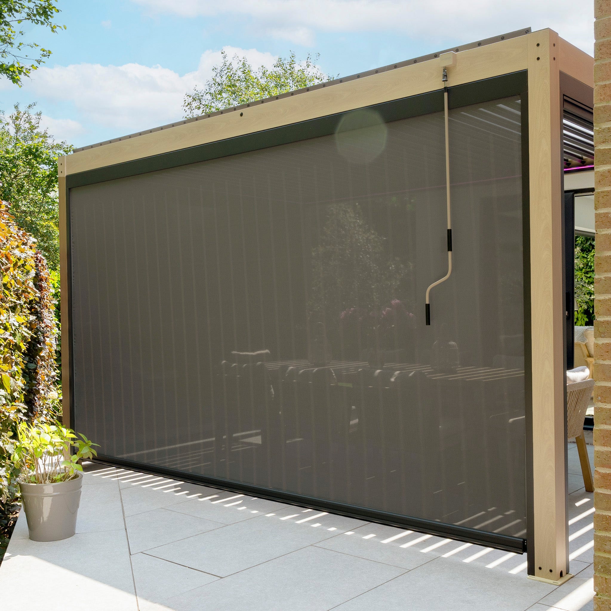 Outdoor patio featuring a beige PergoSTET Plus 3m x 4m pergola with adjustable louvres, three drop sides, LED lighting, and wood effect, surrounded by lush greenery.